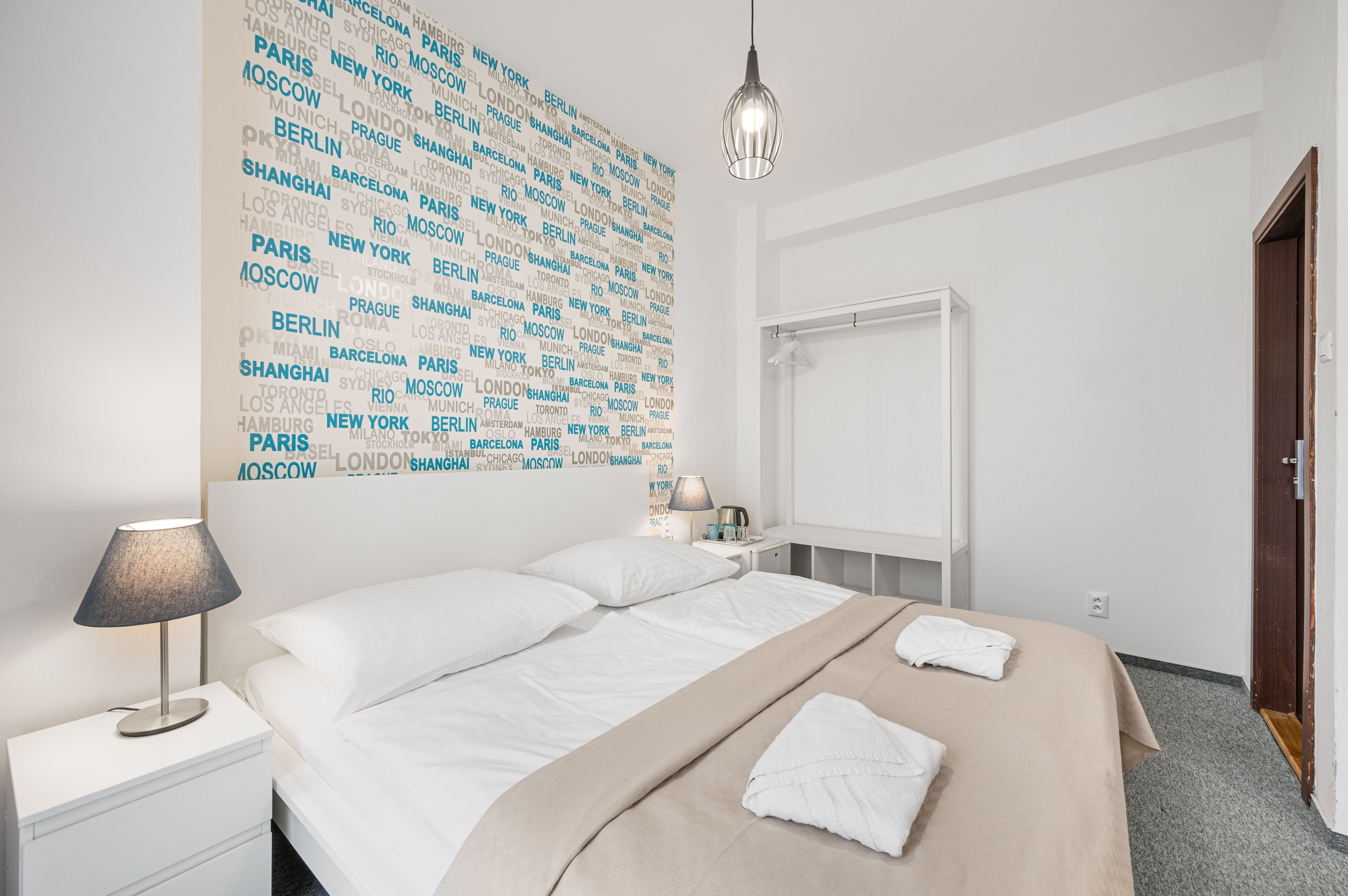 Habitación de hotel con cama, mesita de noche, lámpara, pared con nombres de ciudades y una puerta.