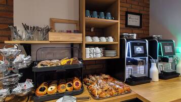 Ein Frühstücksbuffet mit Gebäck, Kaffeeautomaten und einer Schautafel mit Kaffee- und Teeauswahl.