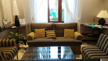 Un salón con un sofá marrón estampado, dos cojines amarillos, una mesa de centro de vidrio, sillones a rayas y una mesita de noche con una lámpara.