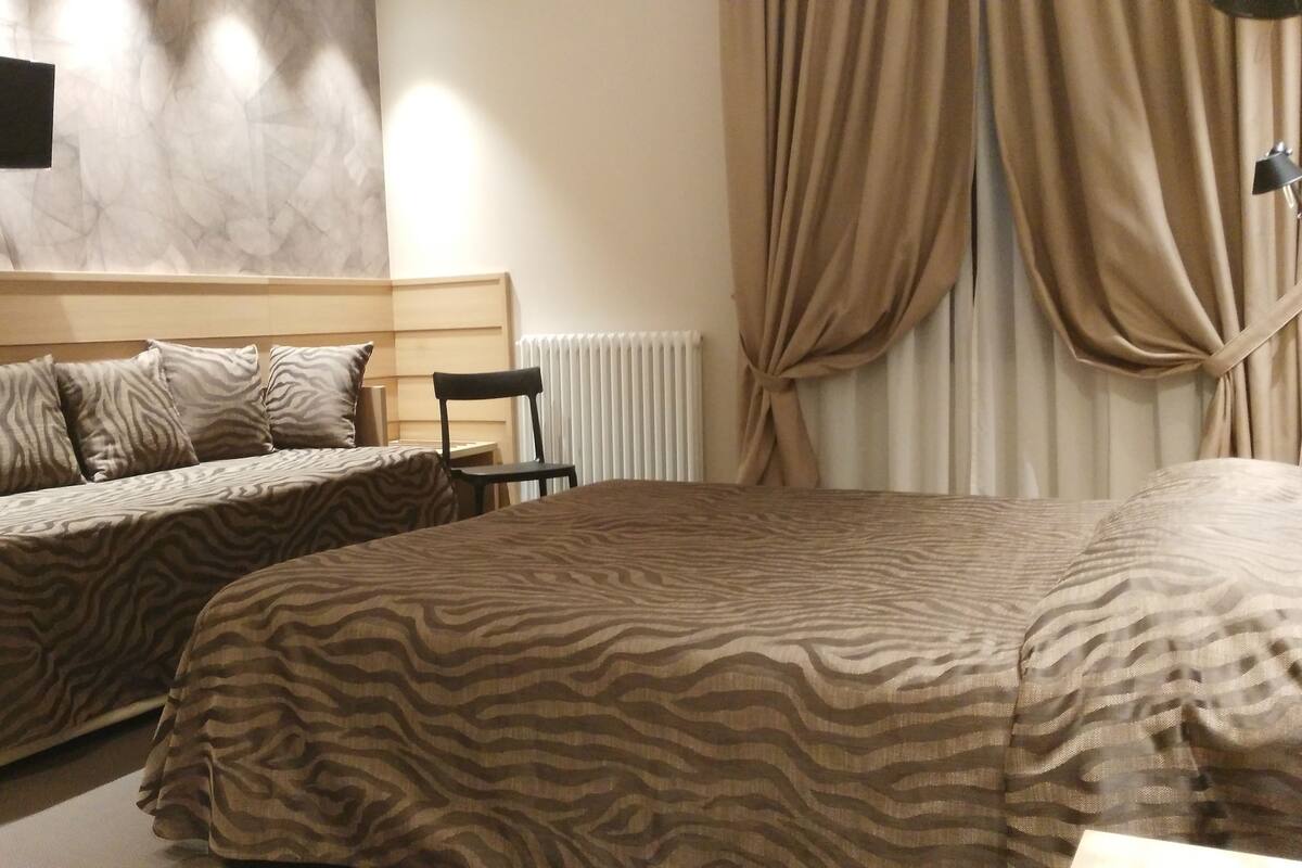 A bedroom with a bed, a chair, a TV mounted on the wall, and a window with curtains.