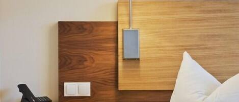 A hotel room with a wooden headboard, a bedside table with a lamp, a phone, and a power outlet.