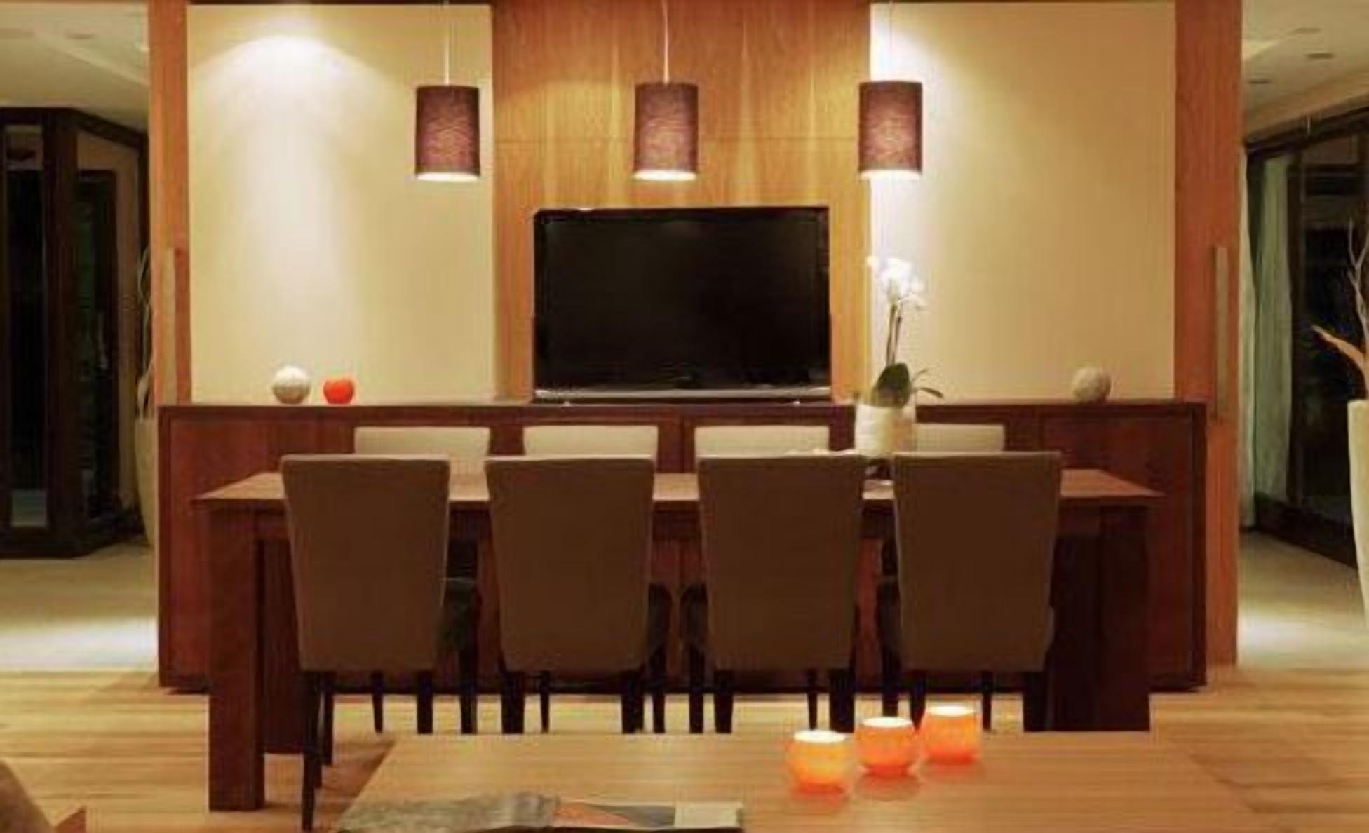 Un comedor moderno con una mesa y sillas de madera, un televisor de pantalla plana, lámparas colgantes y un florero con una planta.