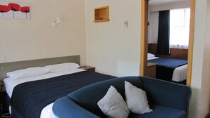 A hotel room with a bed, a chair, a small wooden shelf, and a mirror reflecting the bed.