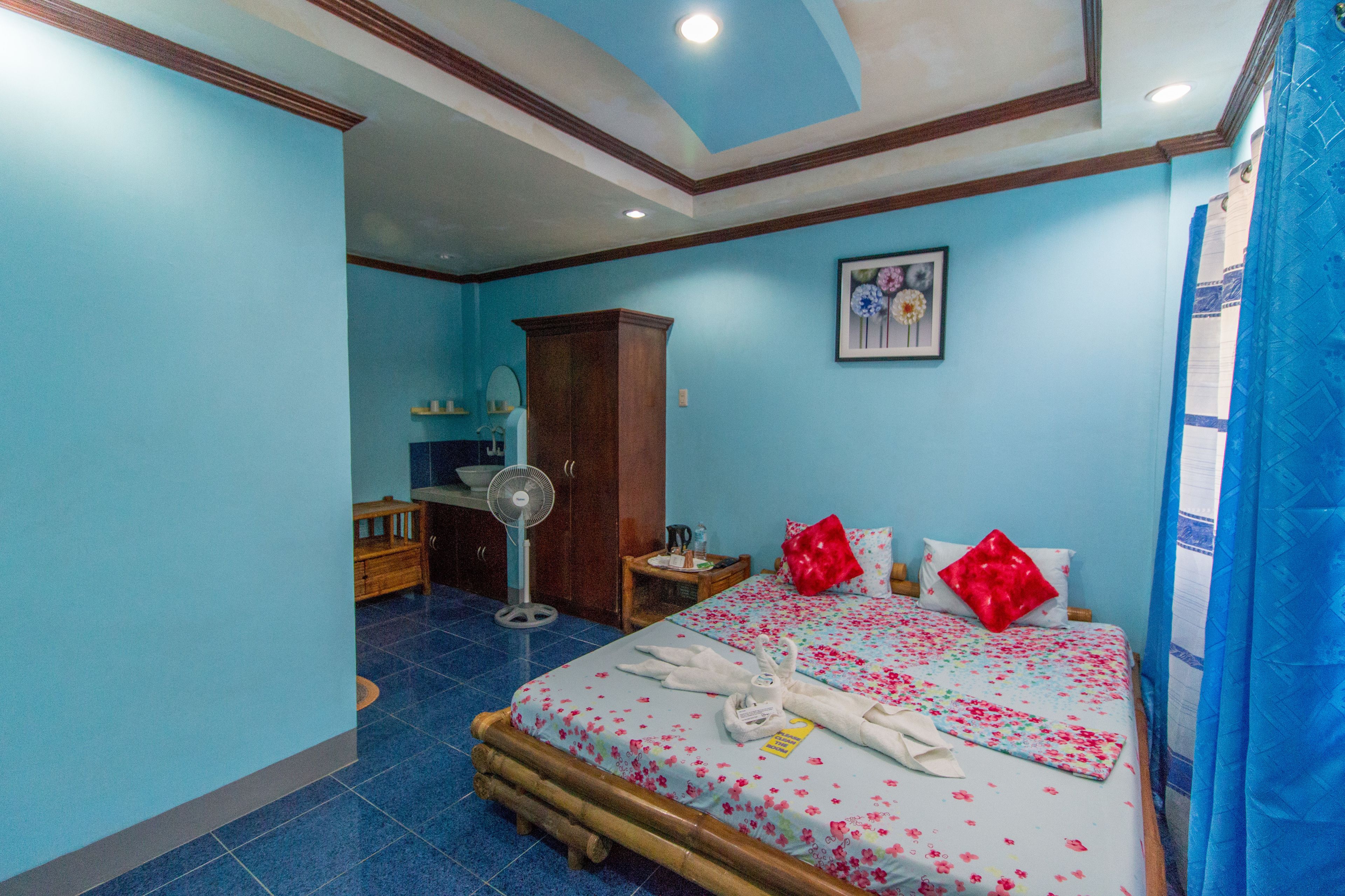 A hotel room with a bed, a wooden wardrobe, a desk, and a fan.