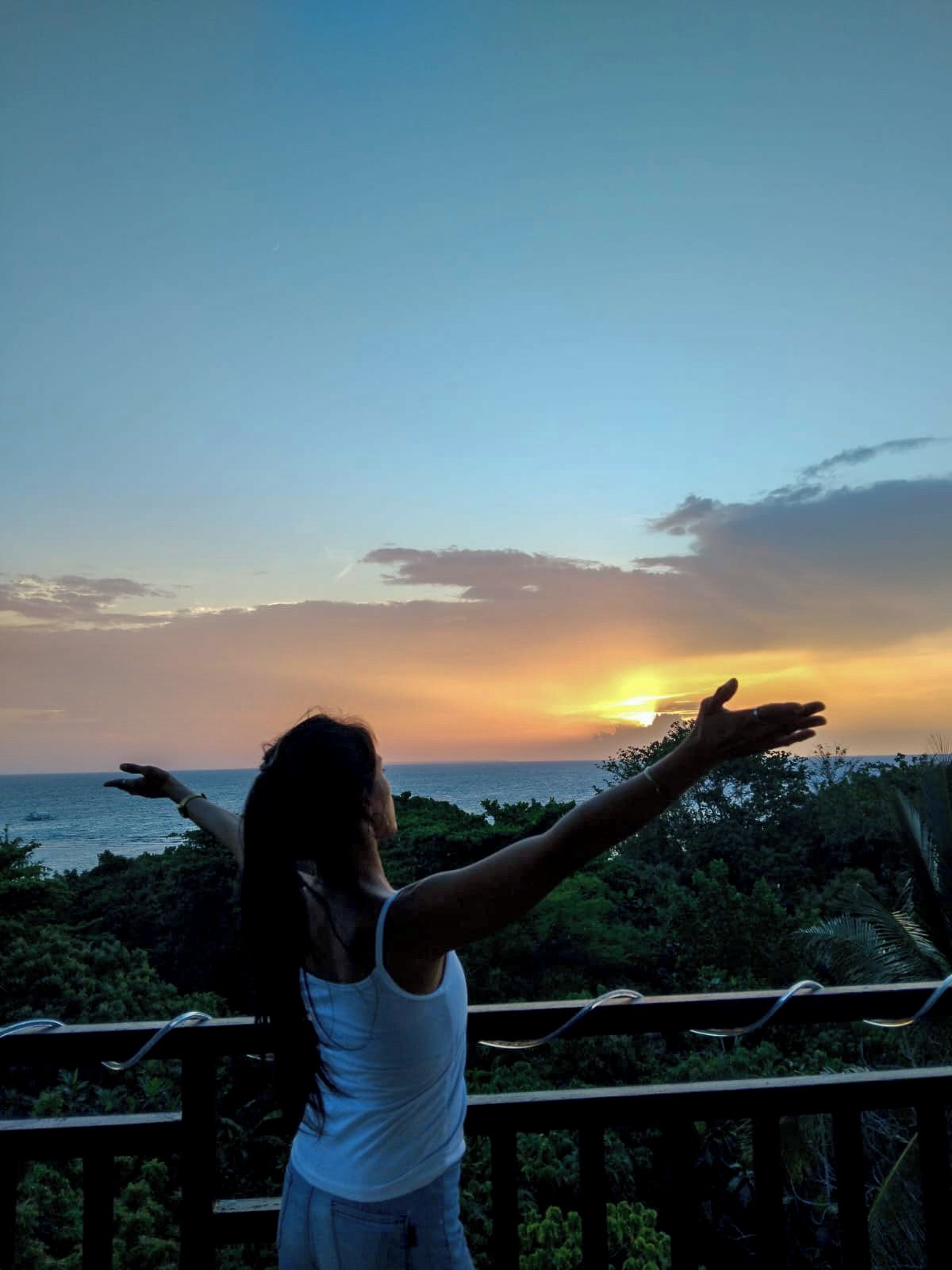Una persona de pie con los brazos extendidos en un balcón, con vistas a un atardecer.