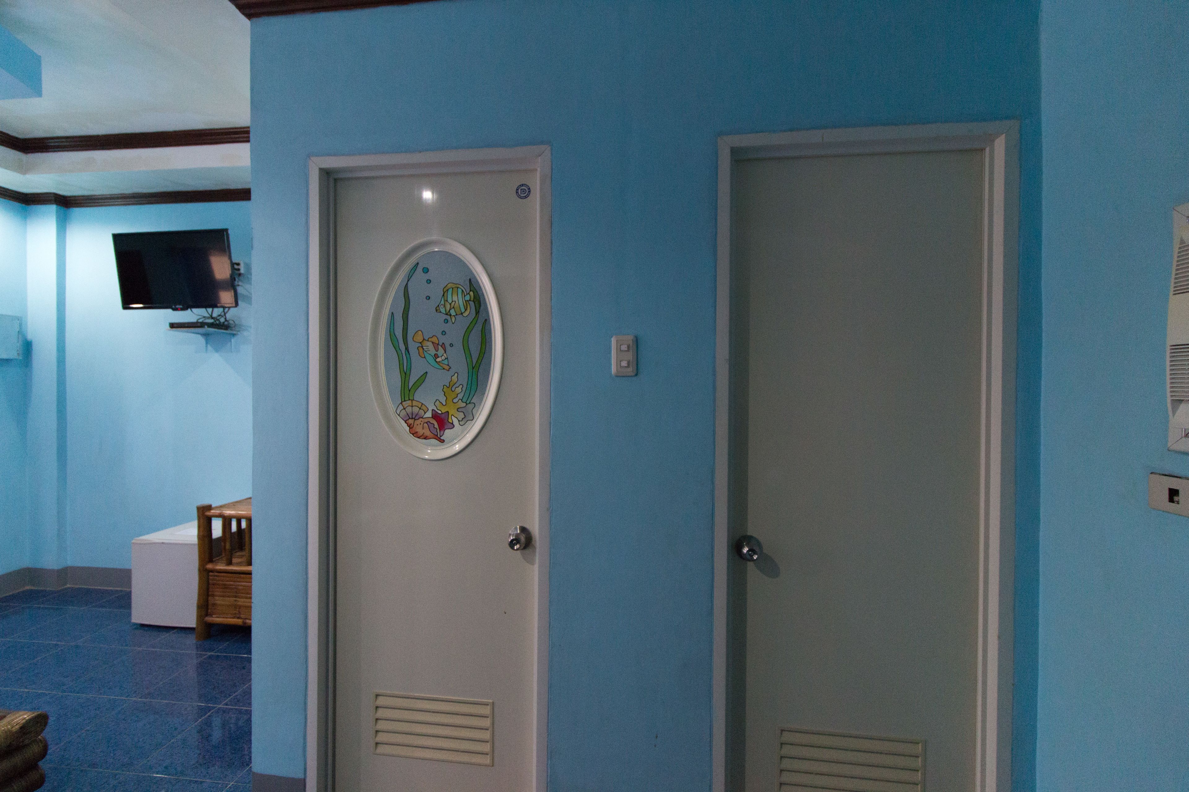 A room with blue walls, a white door with a colorful fish decal, a television mounted on the wall, and a small wooden chair.