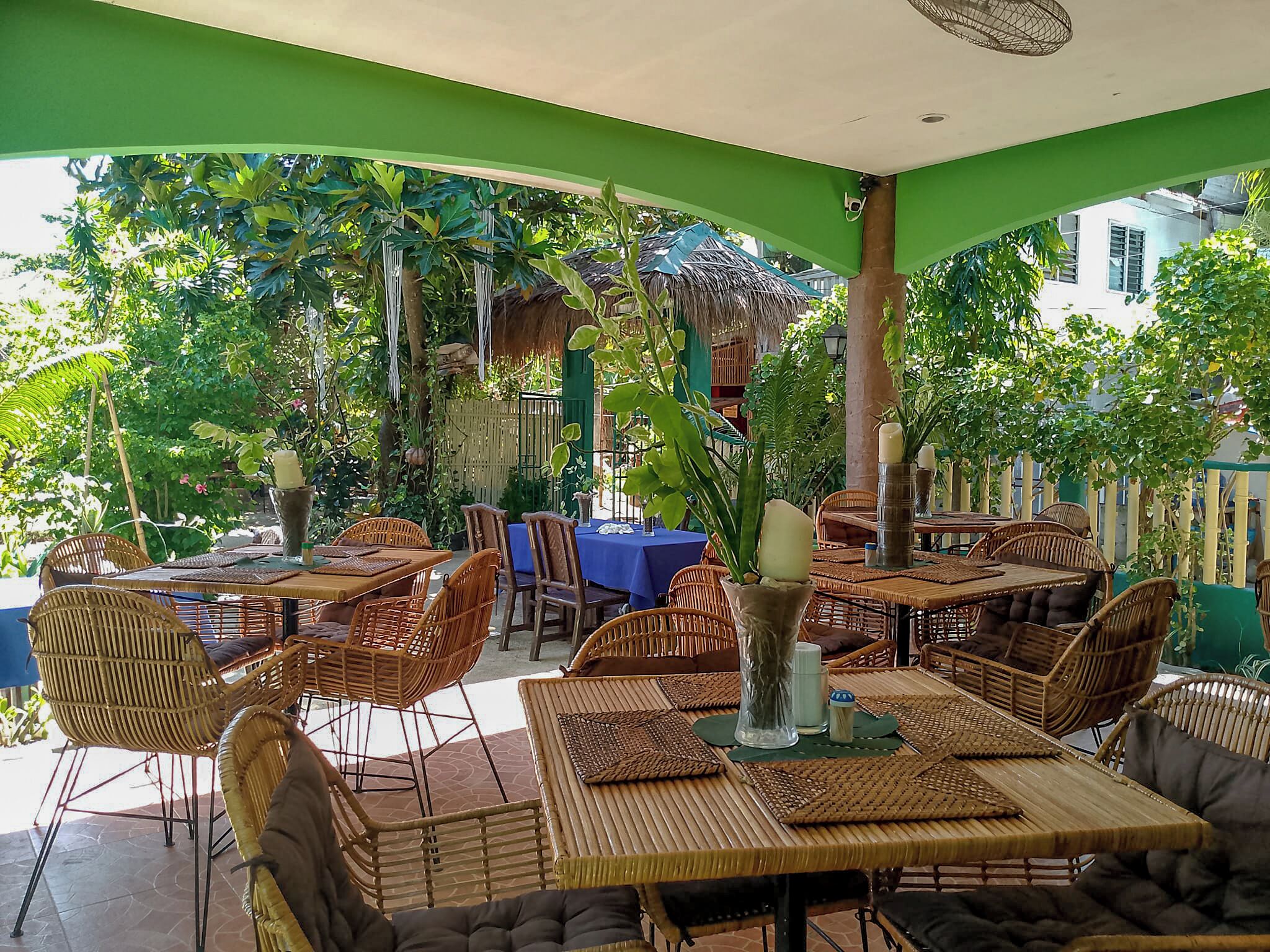 Un área de comedor al aire libre con mobiliario de mimbre, mesas y sillas, rodeada de follaje y una estructura de techo de paja.