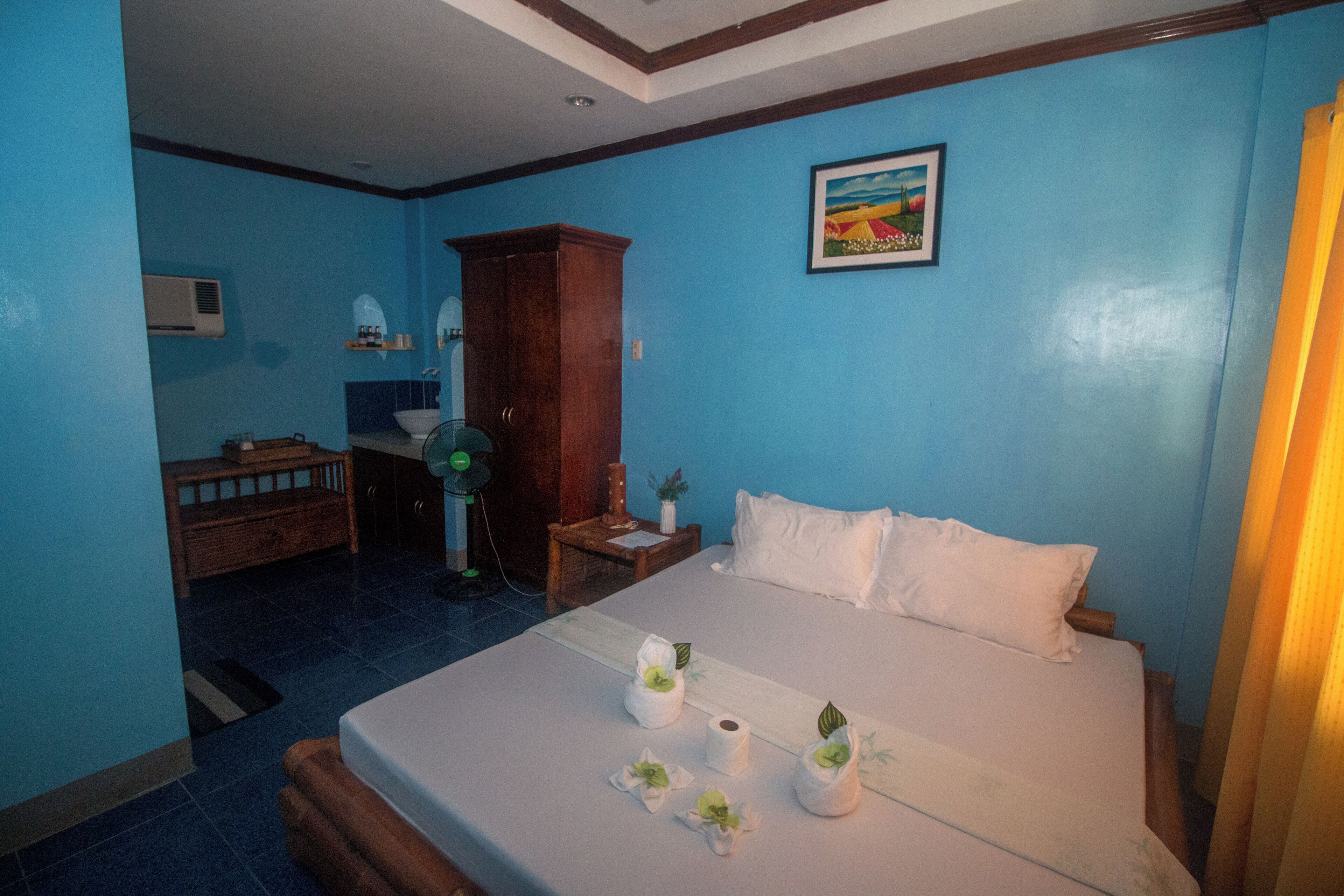 A hotel room with a bed, a wooden cabinet, a small table with a fan, and a picture on the wall.