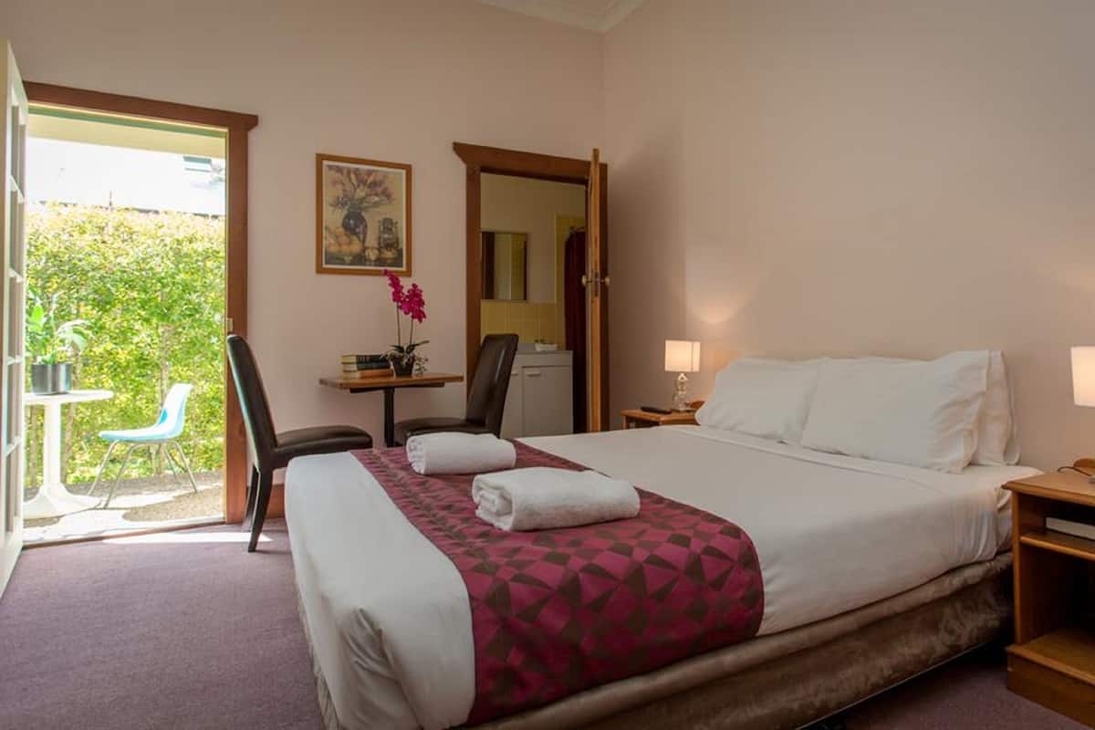 A hotel room with a bed, two bedside lamps, a desk with a chair, a small table with a plant, and a window with a view of greenery.
