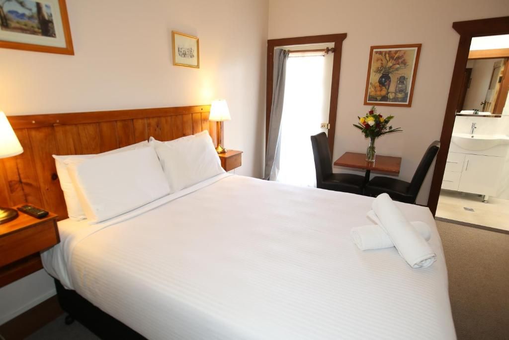 A hotel room with a large bed, bedside lamps, a desk with a chair, a vase of flowers, and a bathroom visible through an open door.