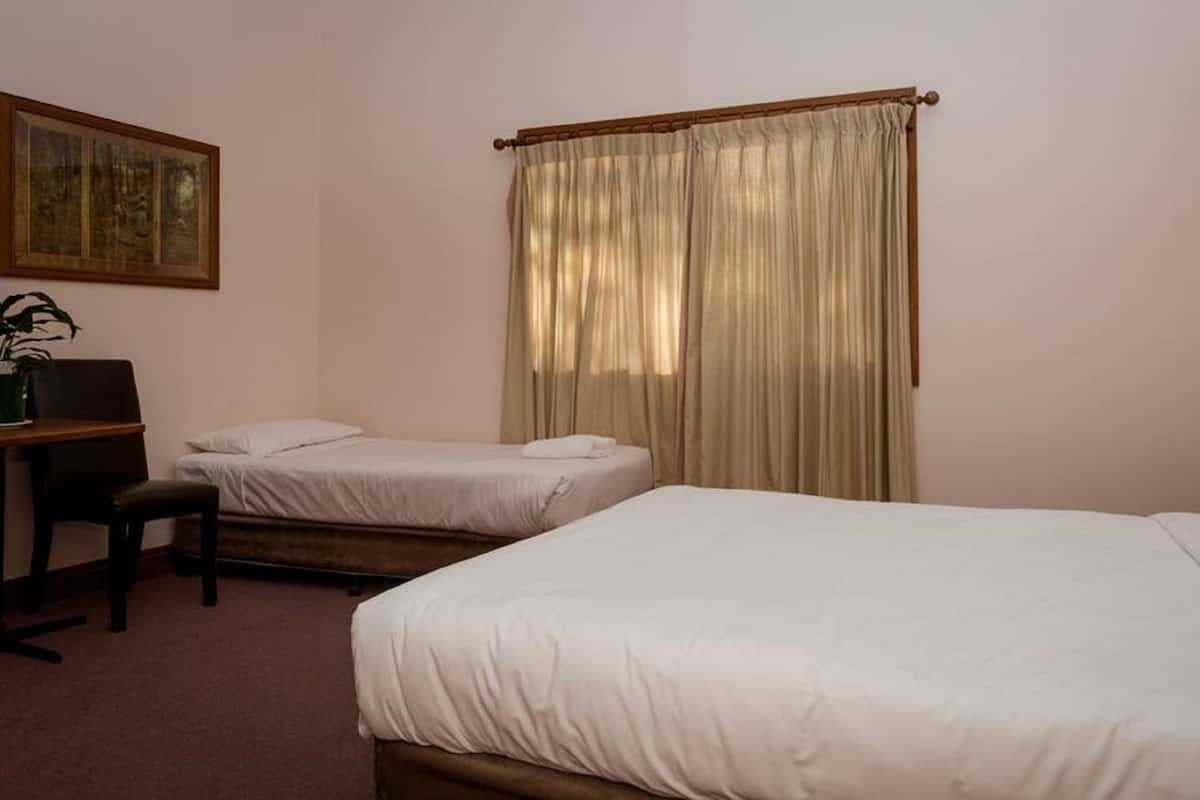 A hotel room with two single beds, a desk with a chair, a lamp, and a large window with curtains.