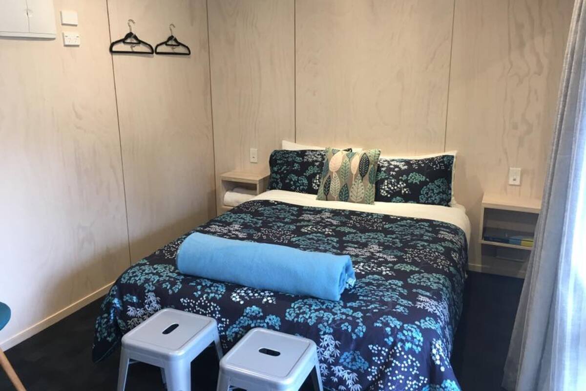 A hotel room with a bed, two nightstands, a small stool, and a wall-mounted shelf.