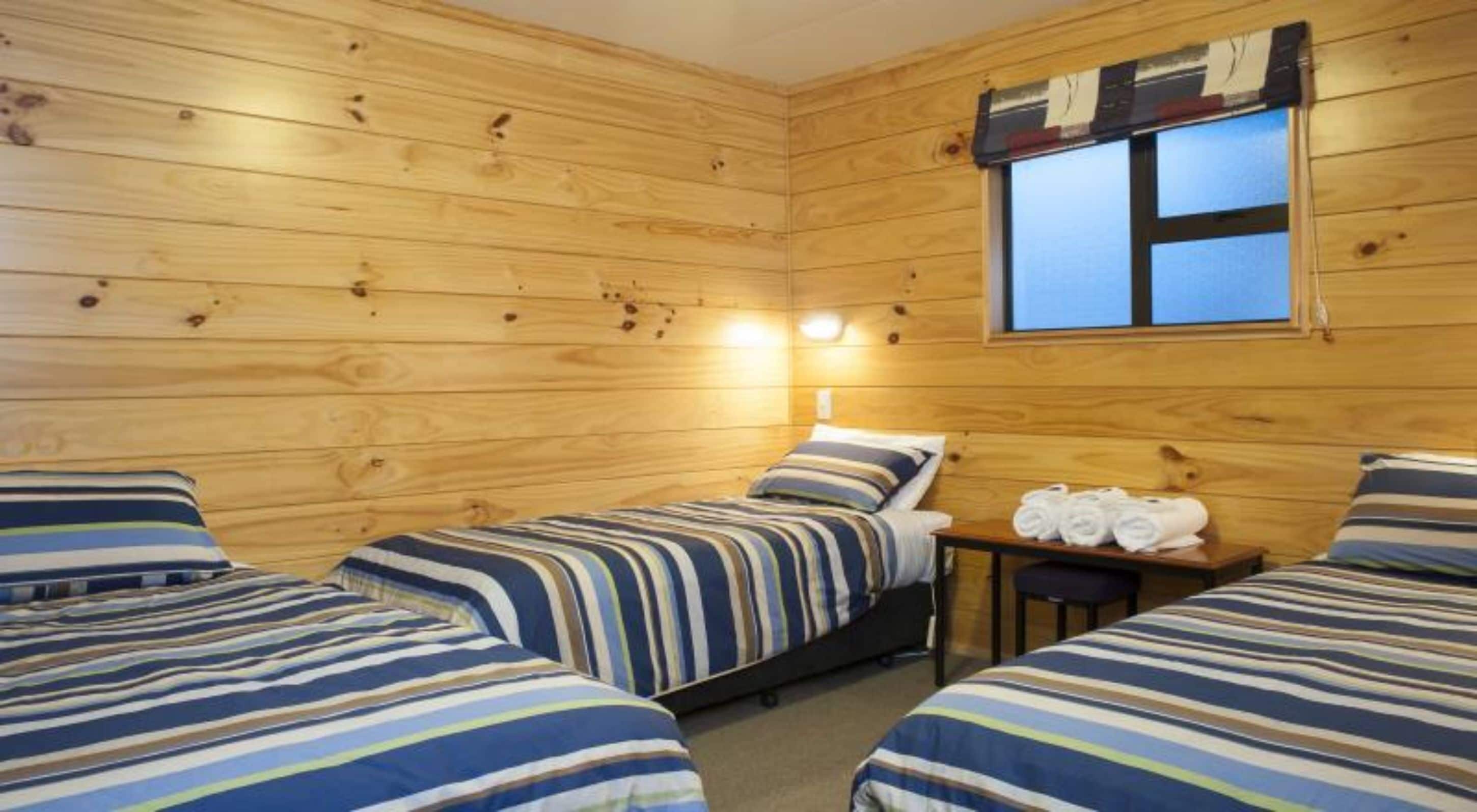 A cabin-style room with two beds, wooden walls, a small table with towels, and a window with curtains.