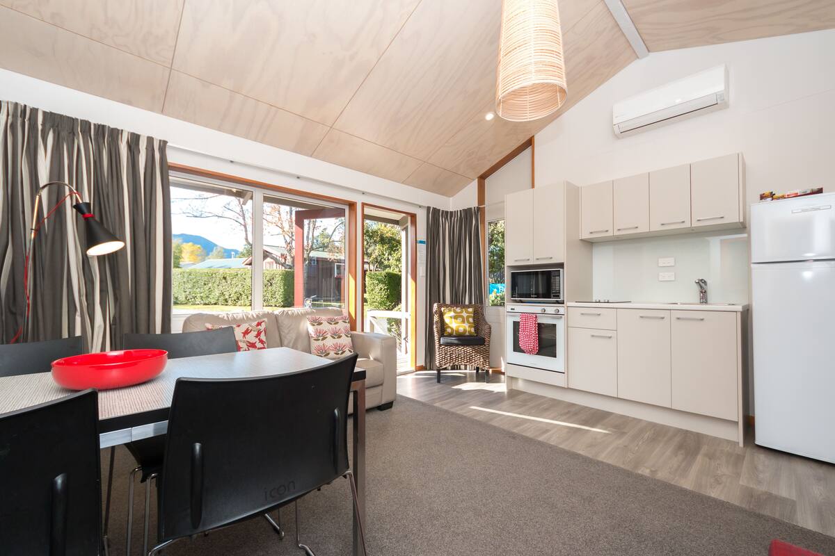 A modern kitchen with white cabinets, a built-in oven, and a refrigerator. There is a dining area with a table and chairs, and a sofa with a patterned cushion.