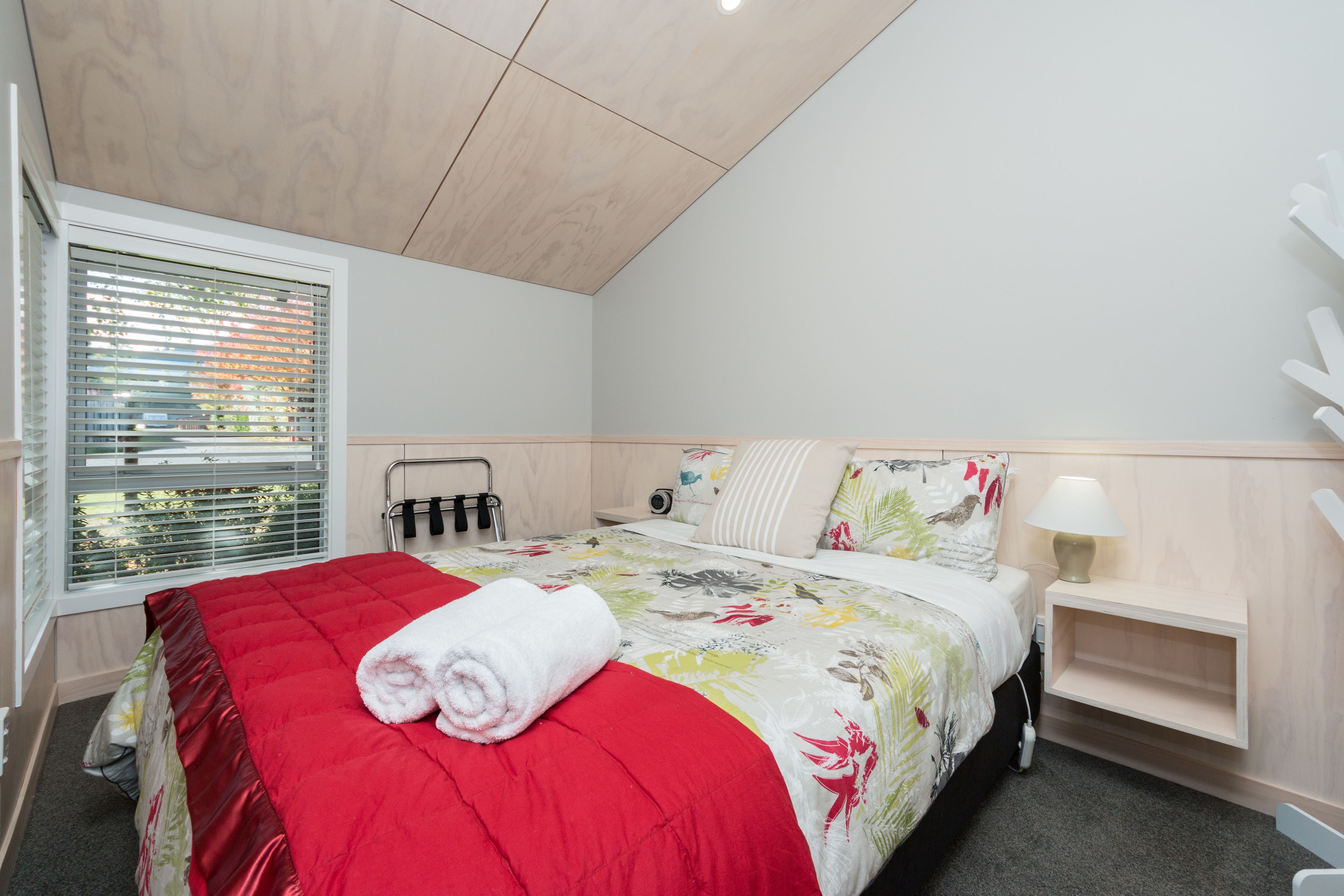 Un dormitorio con cama, mesita de noche con lámpara, ventana con persianas y techo con paneles de madera.