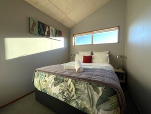 A bedroom with a bed, a nightstand, a lamp, a window, and a painting on the wall. - Hanmer Springs TOP 10 Holiday Park (Hanmer Springs)