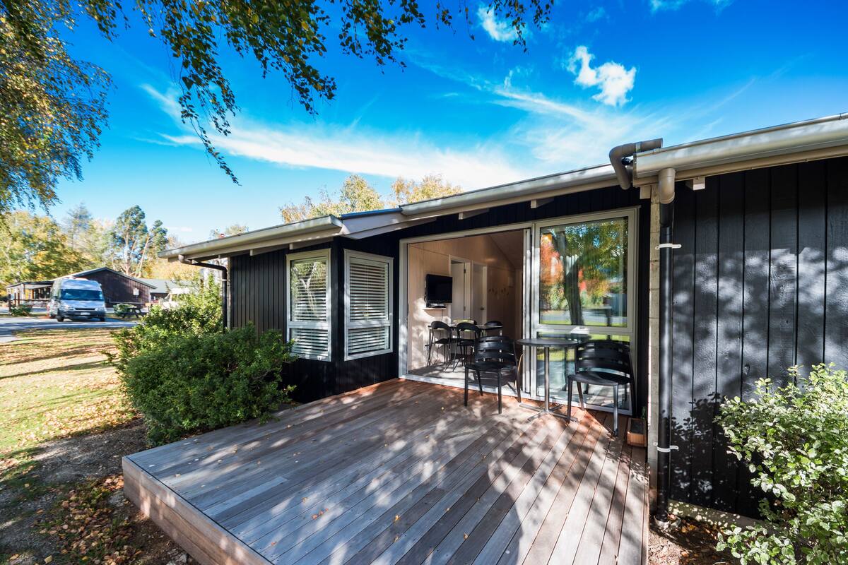 A house with a wooden deck, a sliding glass door leading to a patio with outdoor furniture, and a garage in the background.