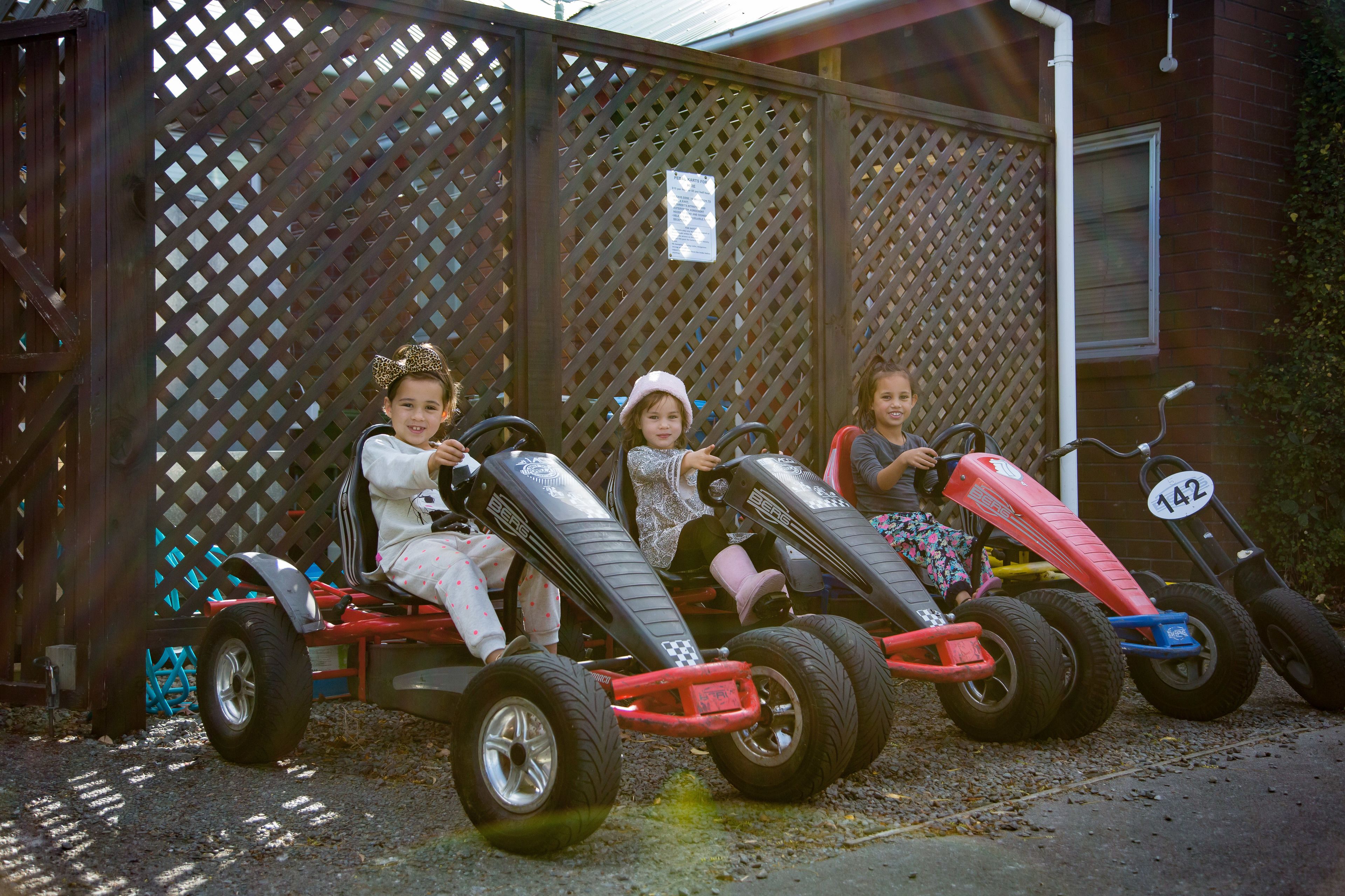 Three children on go-karts parked in front of a wooden fence.