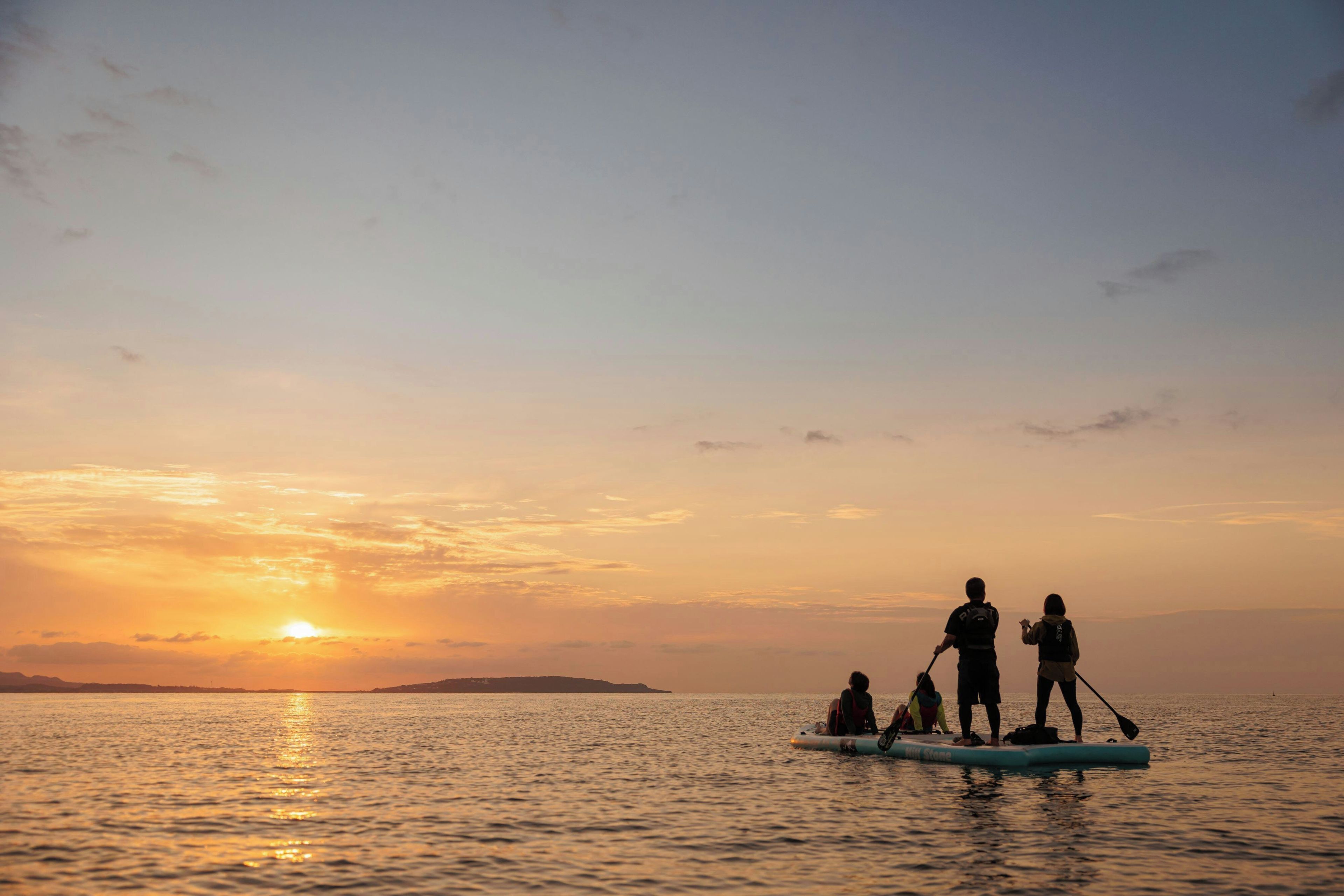 Un coucher de soleil sur une mer calme, avec des personnes sur une planche de paddle.