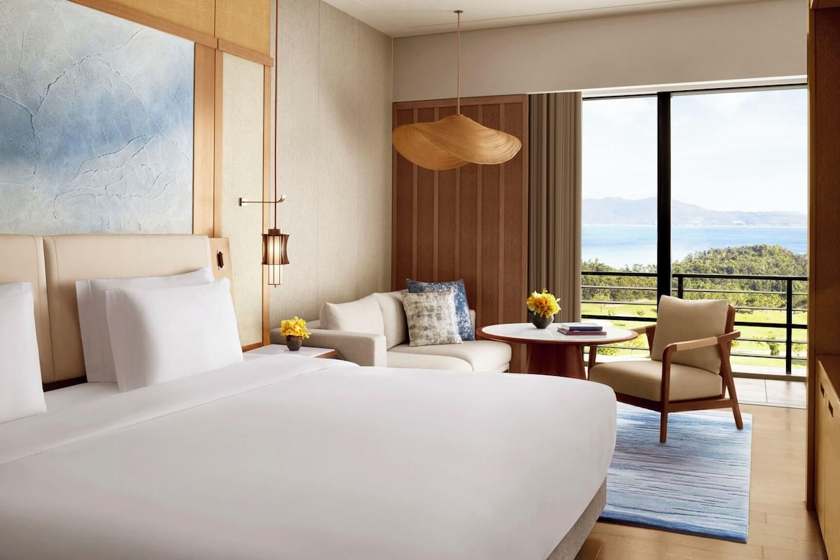 Ein Hotelzimmer mit einem großen Bett, einer Couch, einem kleinen Tisch mit Blumen und Blick ins Freie.