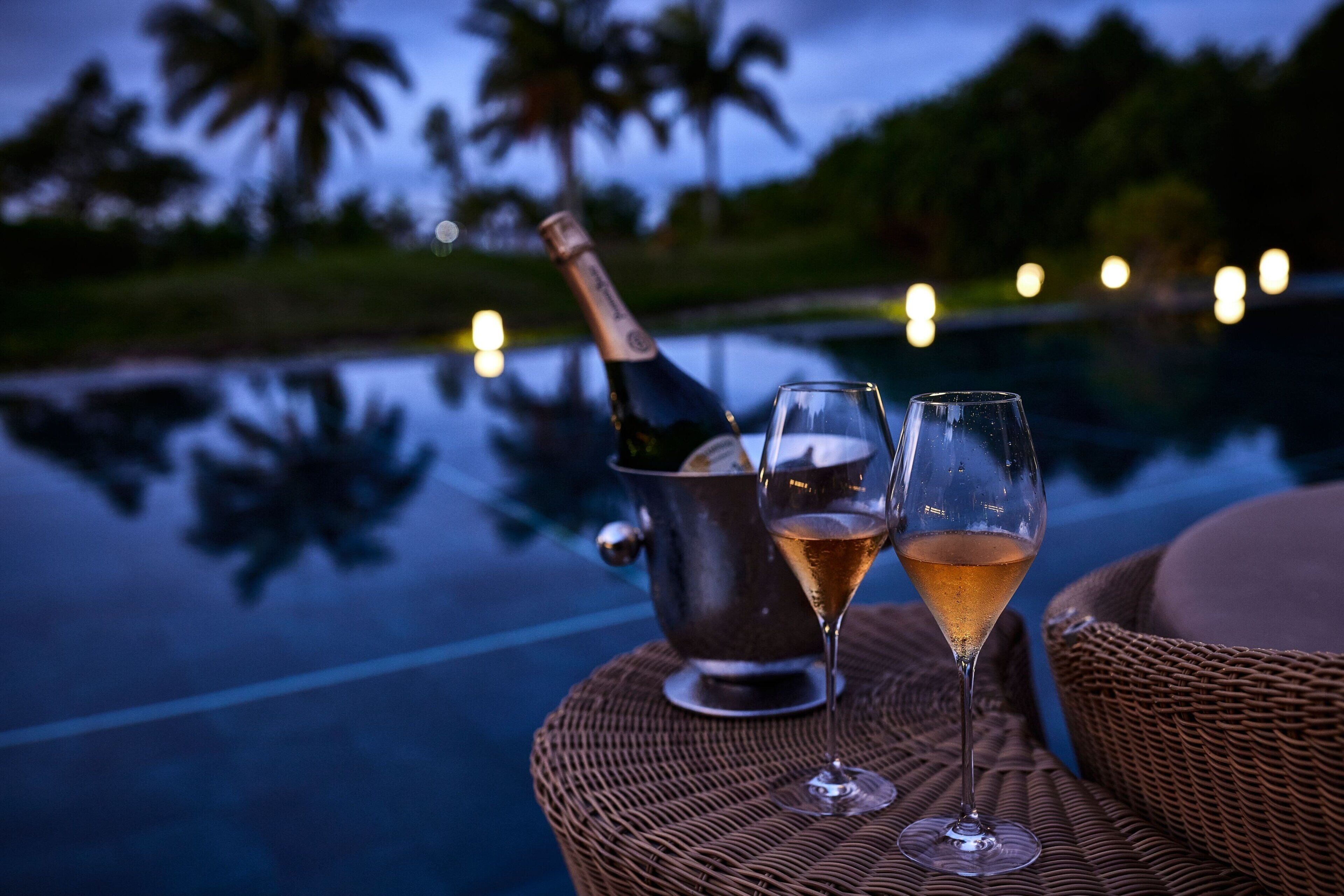A bottle of champagne and two glasses on a wicker table by a pool with palm trees in the background.