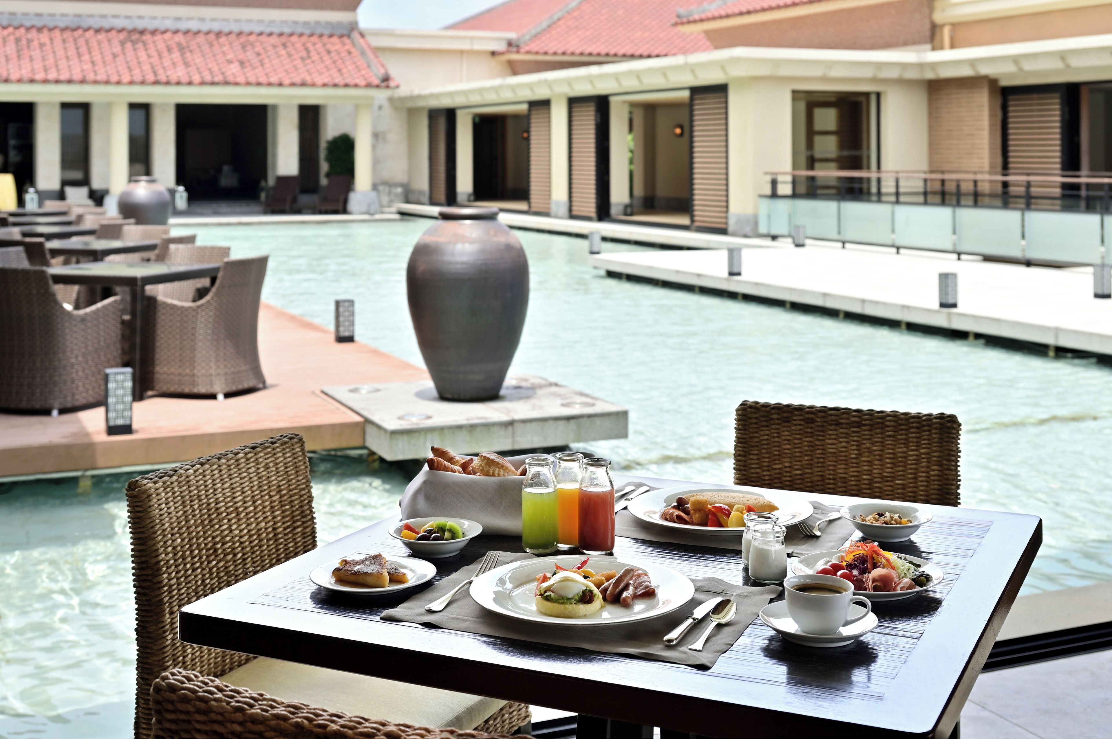 A dining table with breakfast items, a large vase, and a poolside setting.