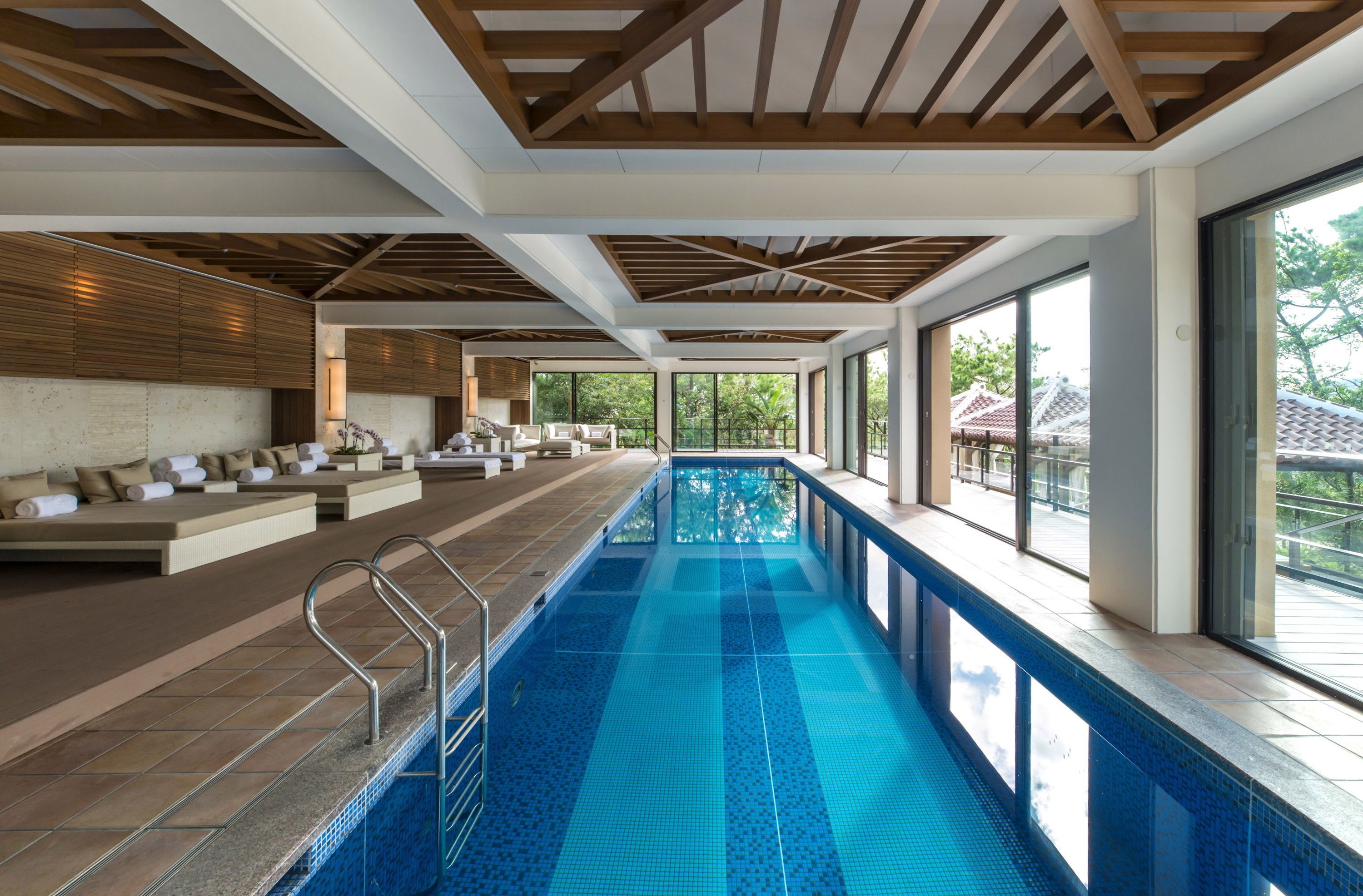 A modern indoor swimming pool with a long bench seating area, large windows, and a wooden ceiling.