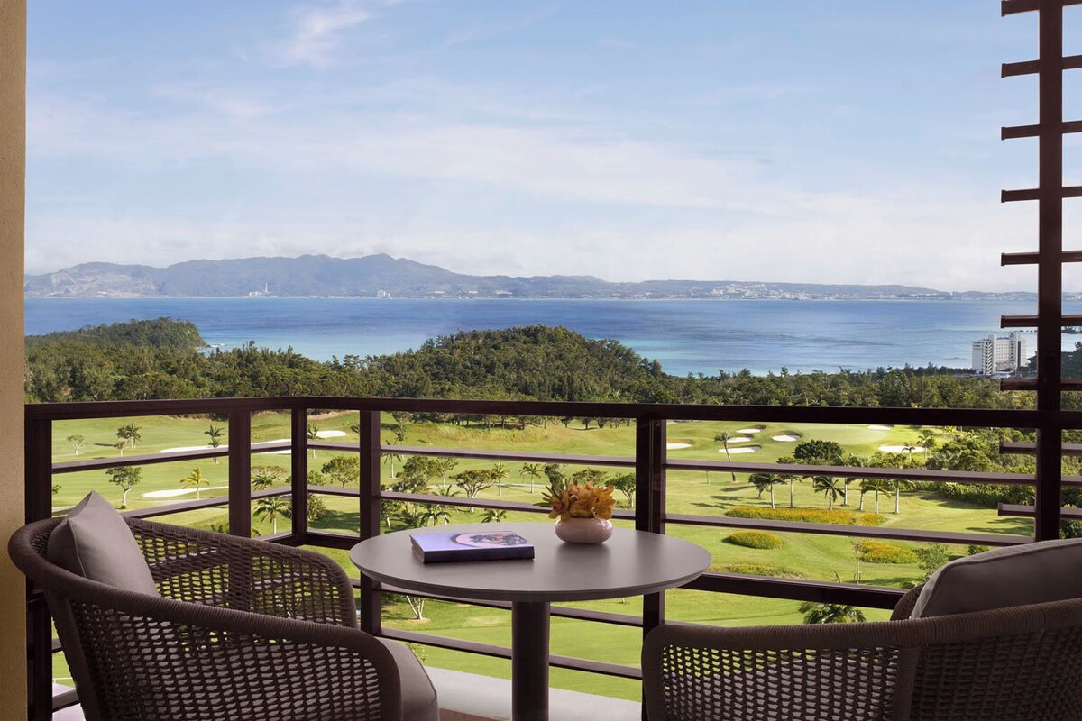 Un balcón con sillas de mimbre y una mesa, con vista a un campo de golf y al mar.