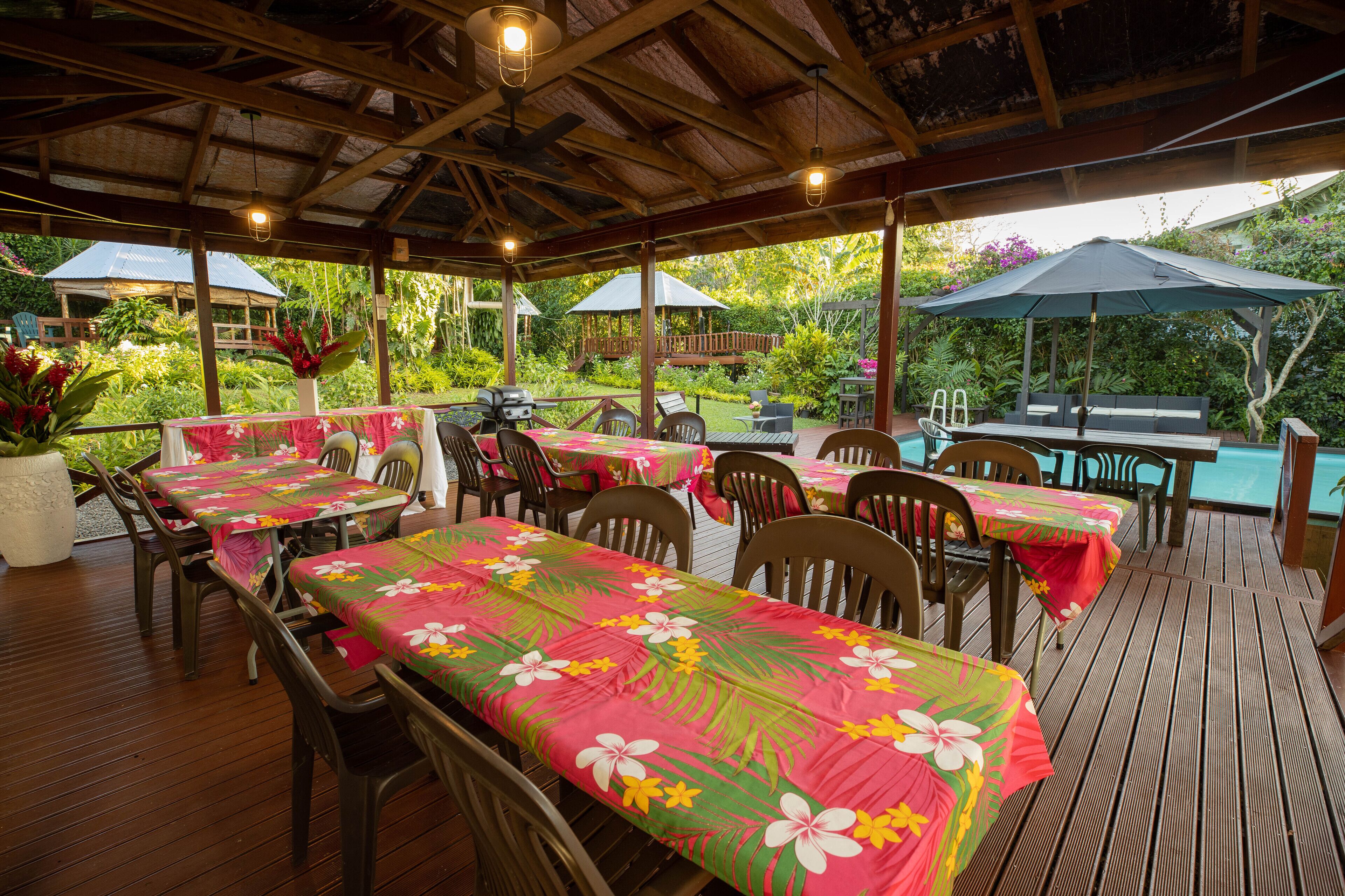 Un área de comedor al aire libre techada, con mesas y sillas, rodeada de abundante vegetación y vista a una piscina.