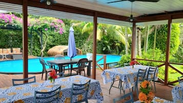 An outdoor pool area with tables and chairs, surrounded by lush greenery and a clear blue pool.