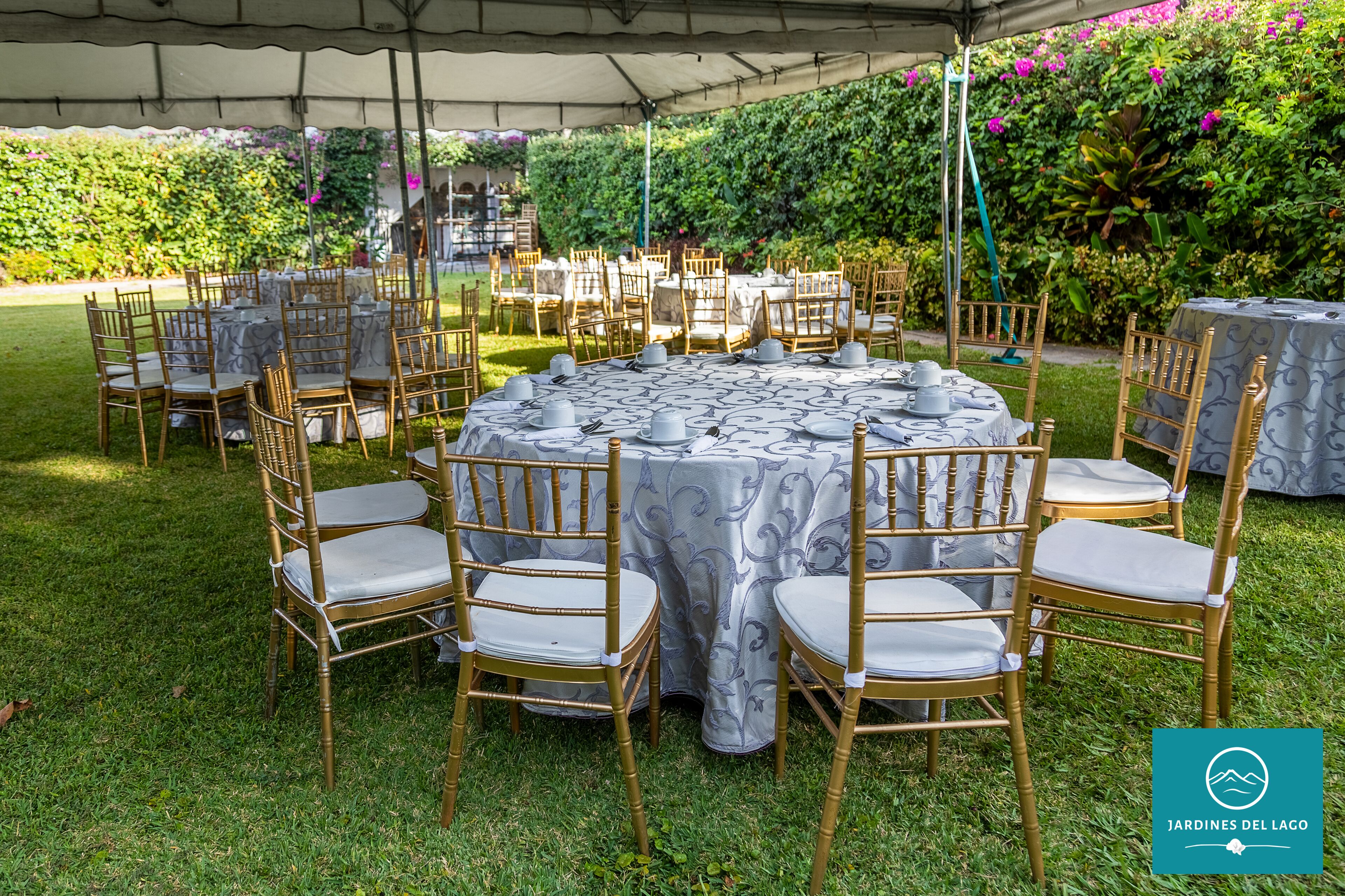 An outdoor tented dining area with round tables and chairs, surrounded by lush greenery.