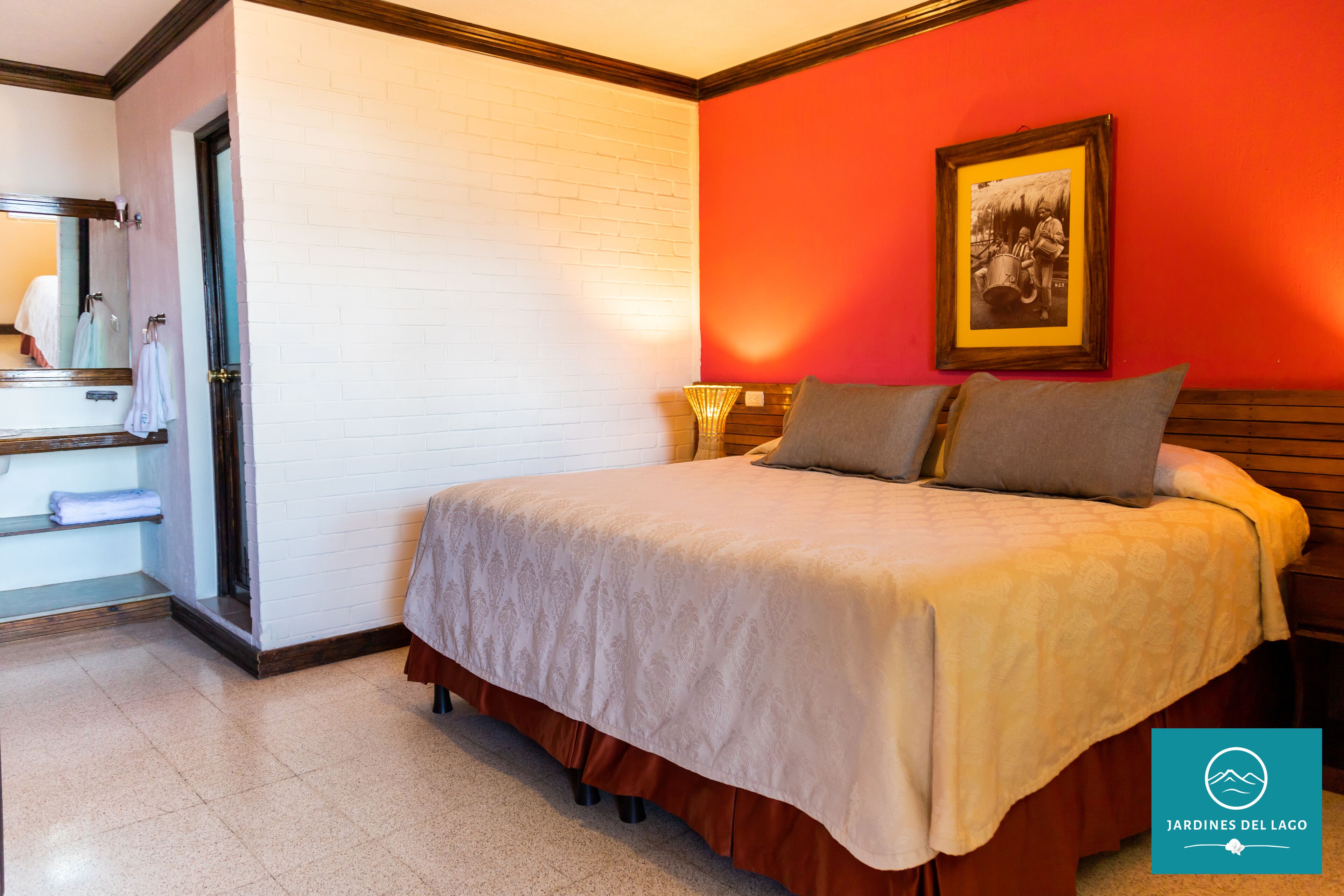 Habitación de hotel con cama, mesita de noche, lámpara, un cuadro en la pared y baño con espejo y toallero.