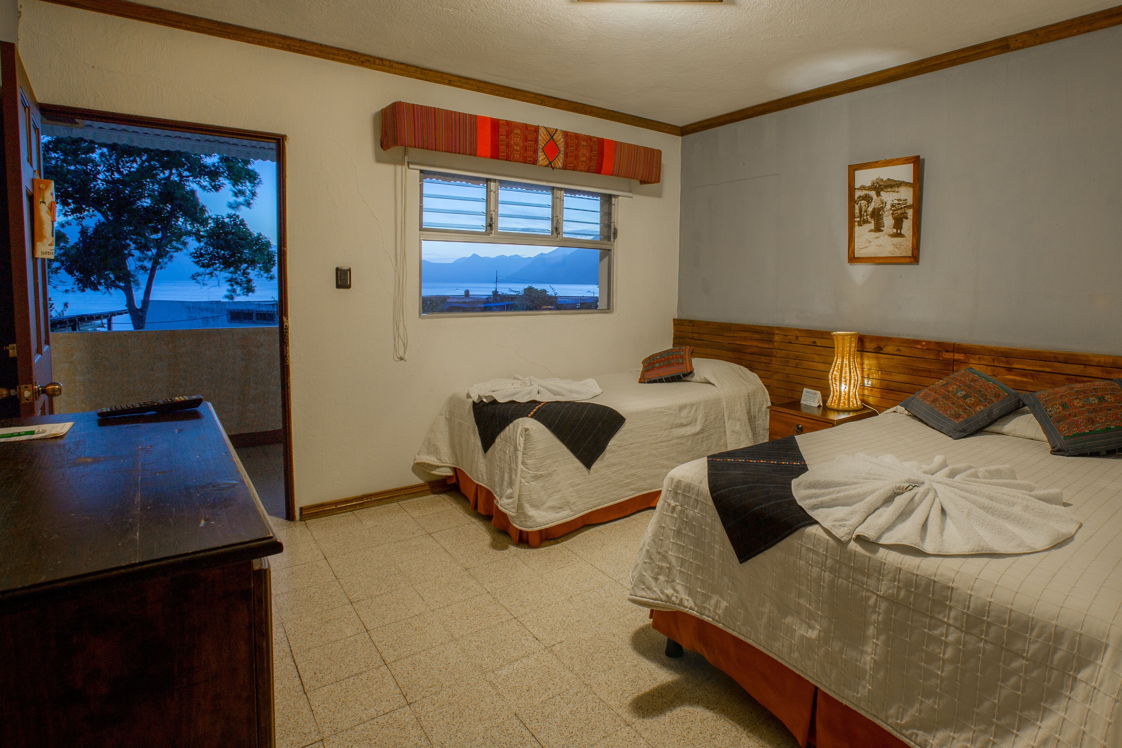 A hotel room with two beds, a desk, a window with a view of a mountain and water, and a framed picture on the wall.