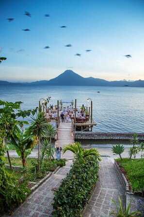 A wooden deck with a stone pathway leading to a seating area by the water, surrounded by lush greenery.