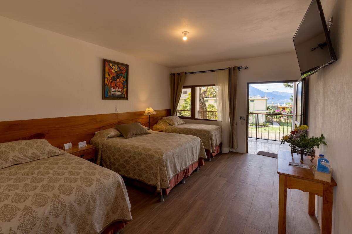 Quarto de hotel com três camas, televisão fixada na parede, janela com cortinas e varanda com vista para as montanhas.