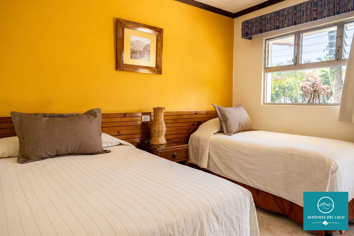 Quarto de hotel com duas camas, cabeceira de madeira, quadro na parede e janela com vista para árvores.