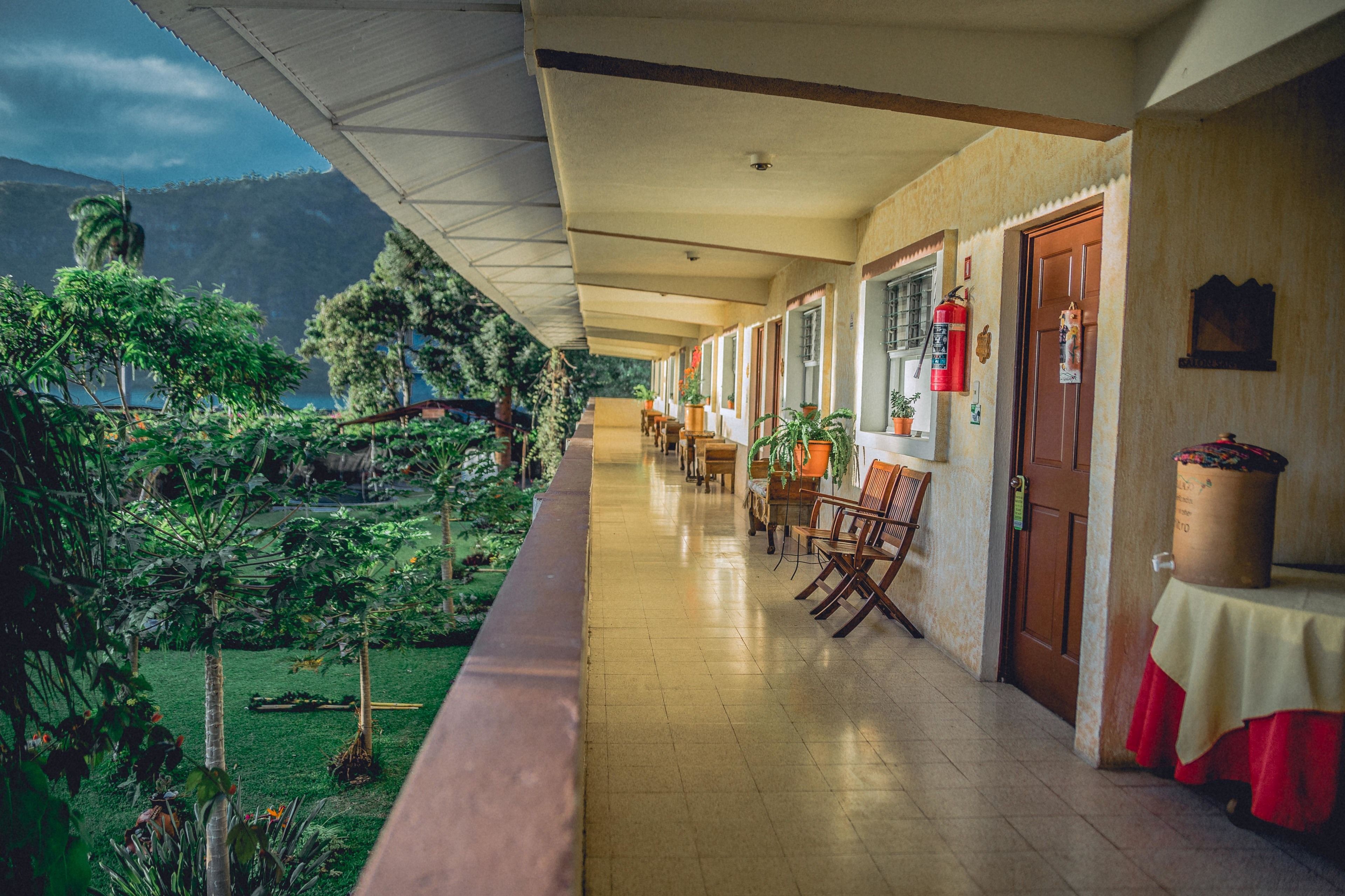 A hotel corridor with a view of a lush garden and mountains. There are wooden chairs and tables along the corridor.