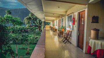 A hotel corridor with a view of a lush garden and mountains. There are wooden chairs and tables along the corridor.