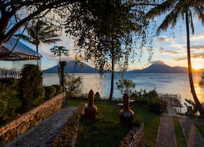 Un jardín con un muro de piedra, un jarrón grande y vista a un lago con montañas al fondo.