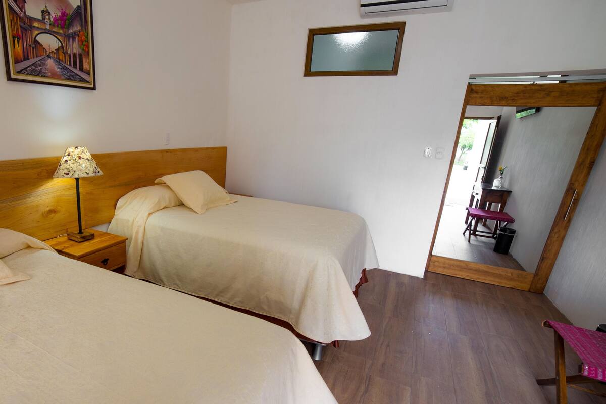 Quarto de hotel com duas camas, uma mesa de cabeceira com abajur, um espelho e piso de madeira.