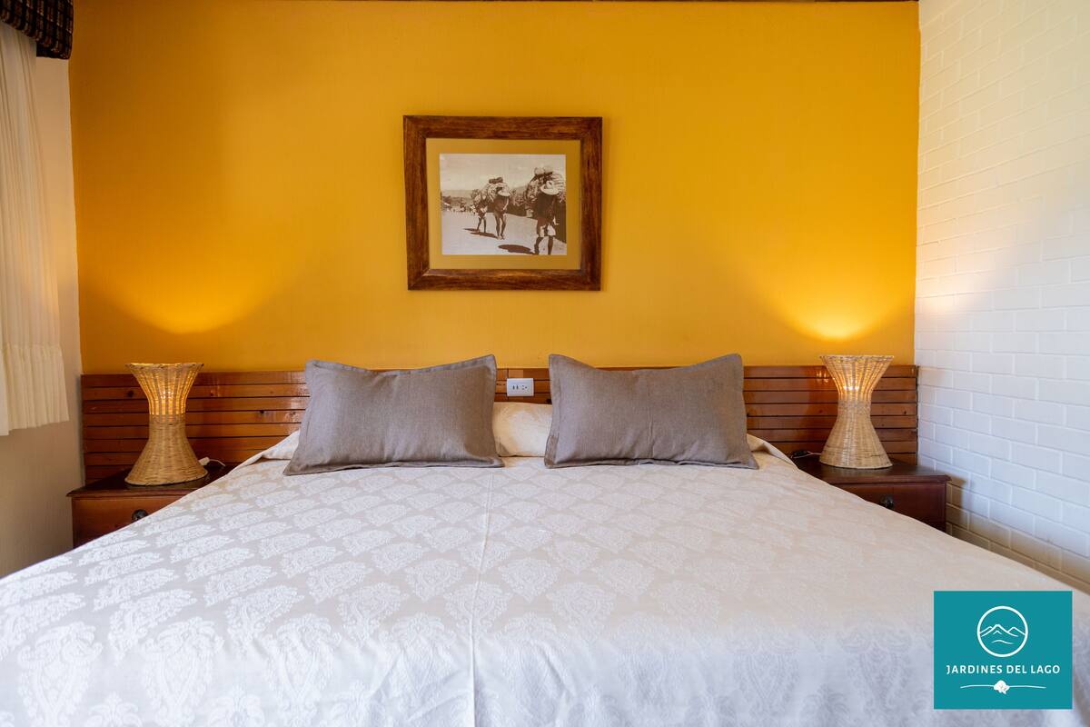 Quarto de hotel com uma cama grande, duas luminárias de cabeceira, um quadro na parede e um logotipo no canto.