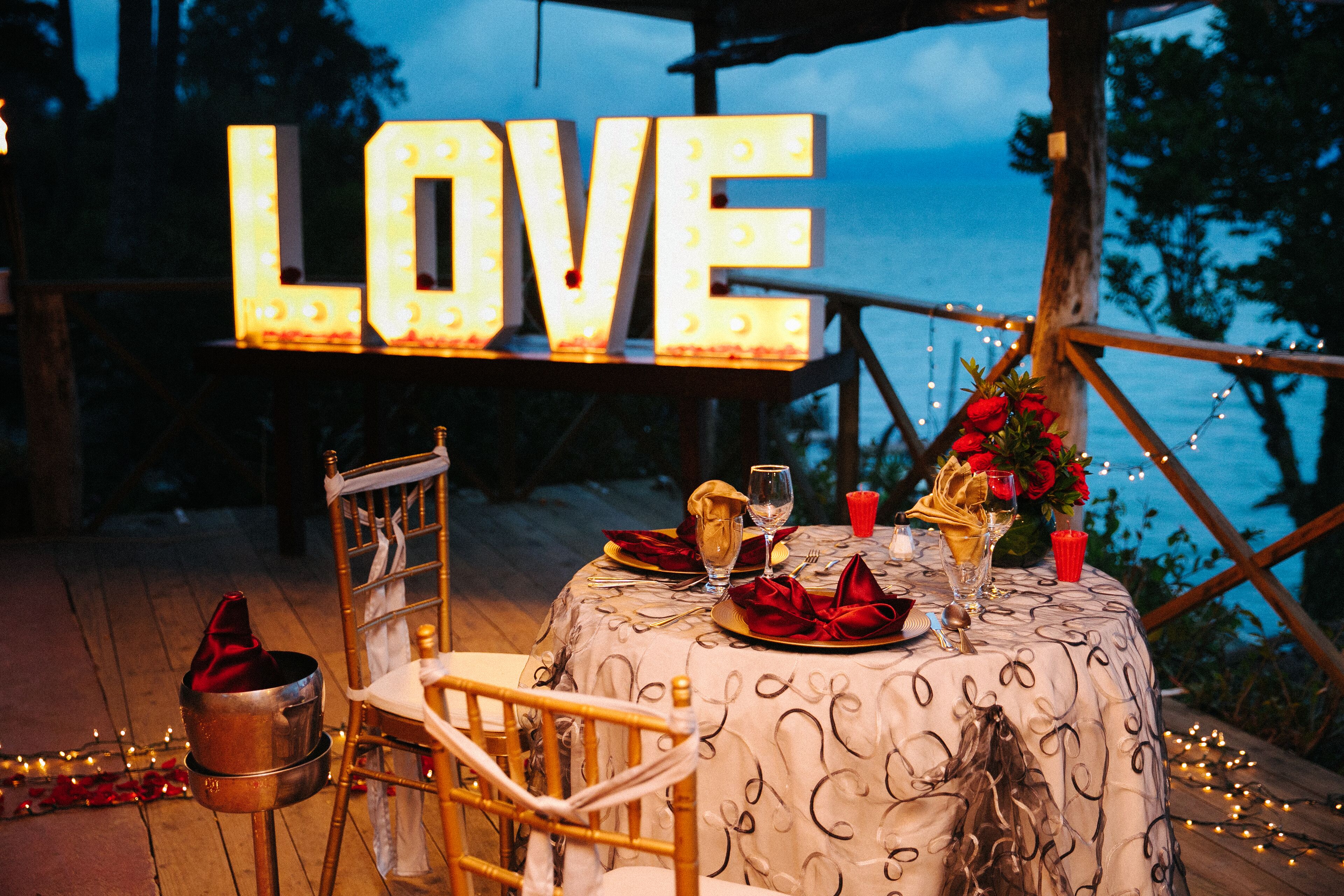 Ein romantisches Essensarrangement im Freien mit einem 'LOVE'-Schild, Tisch gedeckt mit roten Servietten und Blick aufs Meer.