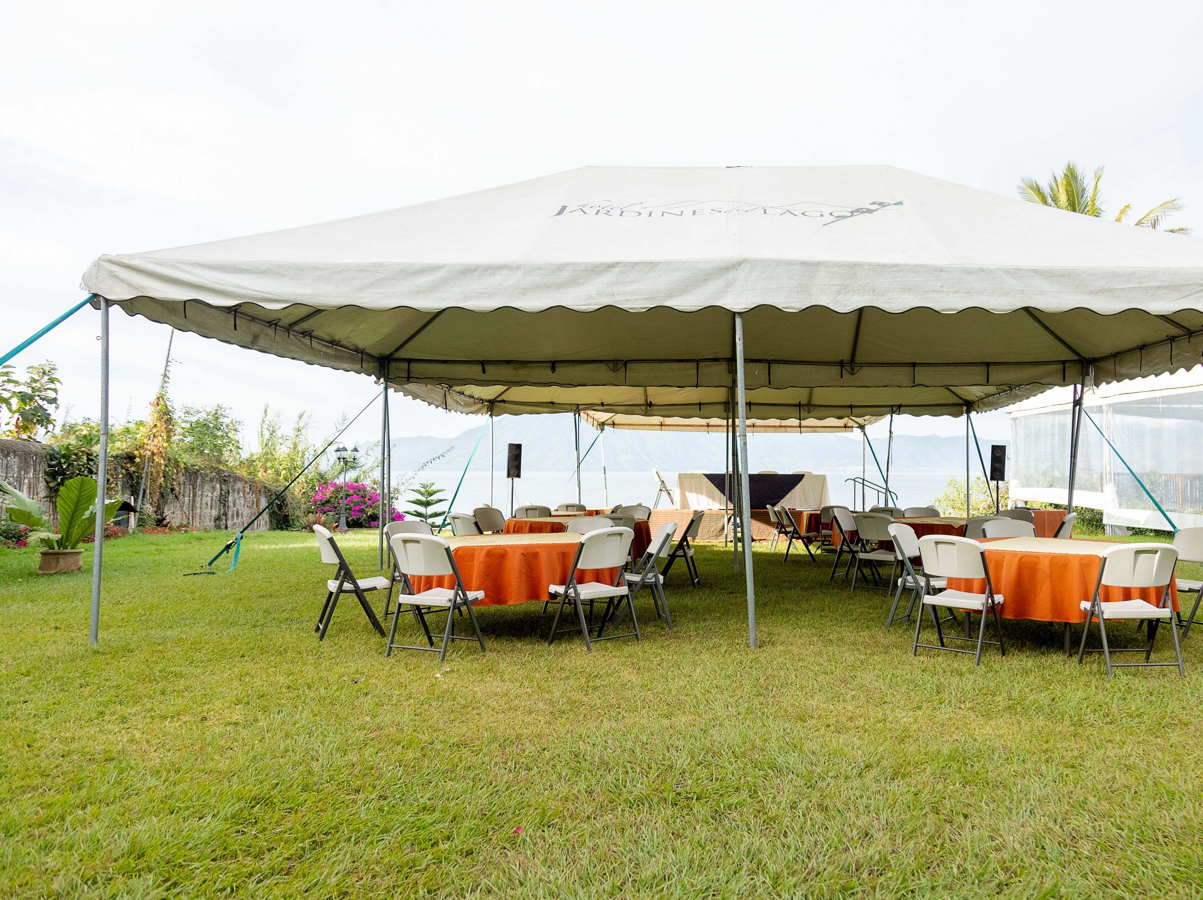 A tented outdoor dining area with tables and chairs, set against a backdrop of a grassy field and a body of water.