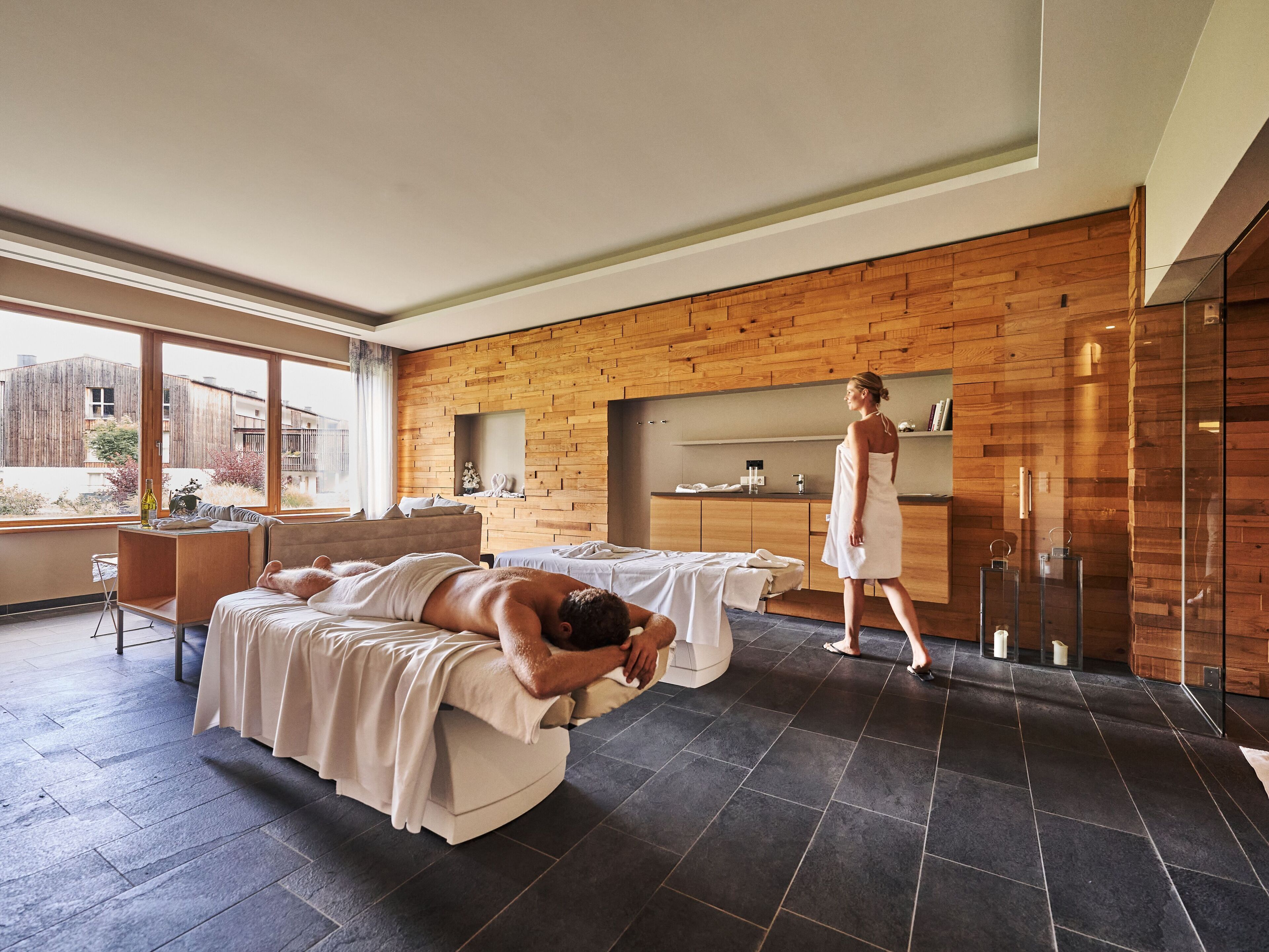 Un cuarto de spa con dos camillas de masaje, una pared de madera, un ventanal grande y una zona de recepción.