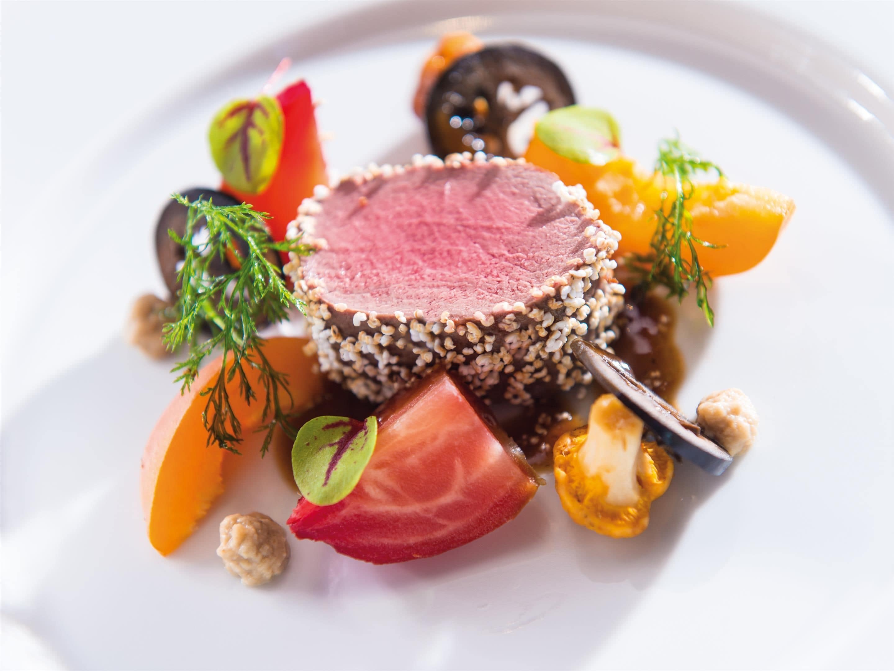 A dish with a slice of meat topped with sesame seeds, surrounded by various vegetables and garnishes.