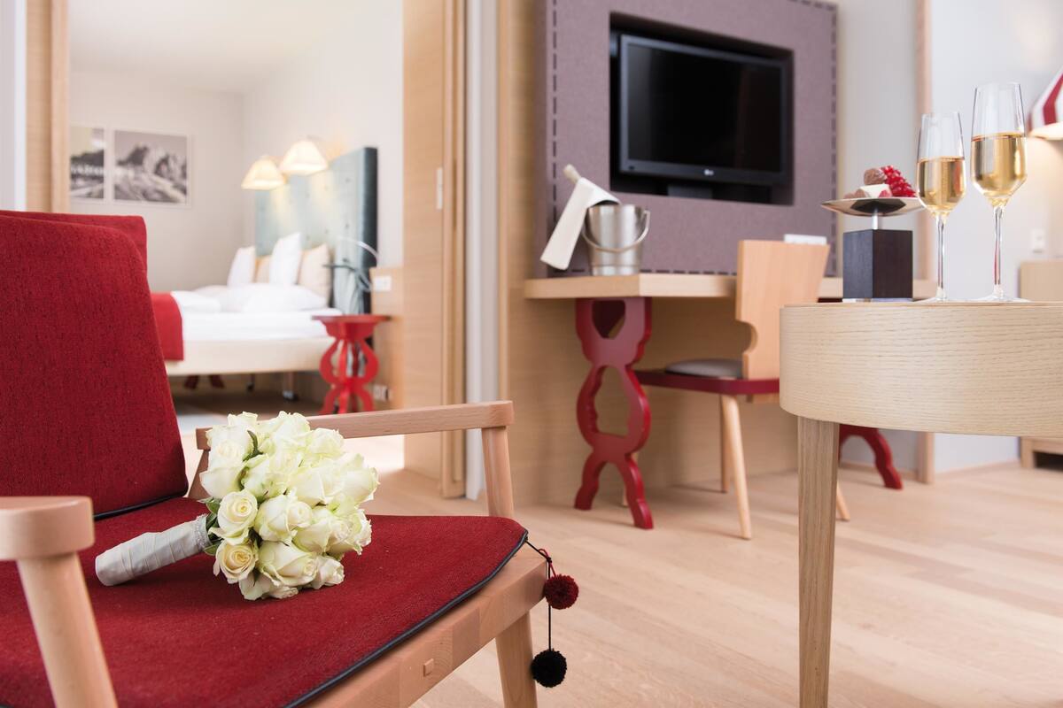 A hotel room with a red chair, a bouquet of white flowers, a wooden desk with a television, and two glasses of champagne.