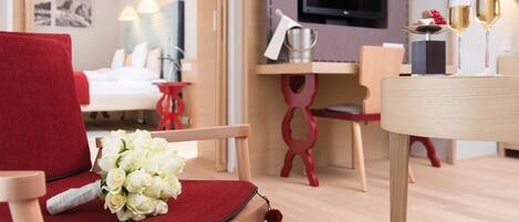A hotel room with a red chair, a bouquet of white flowers, a wooden desk with a television, and two glasses of champagne.