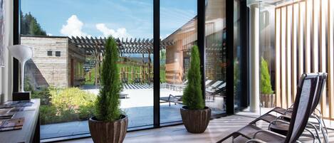 Un interior moderno con grandes ventanales, un patio con mobiliario y vistas a un edificio de madera y vegetación.