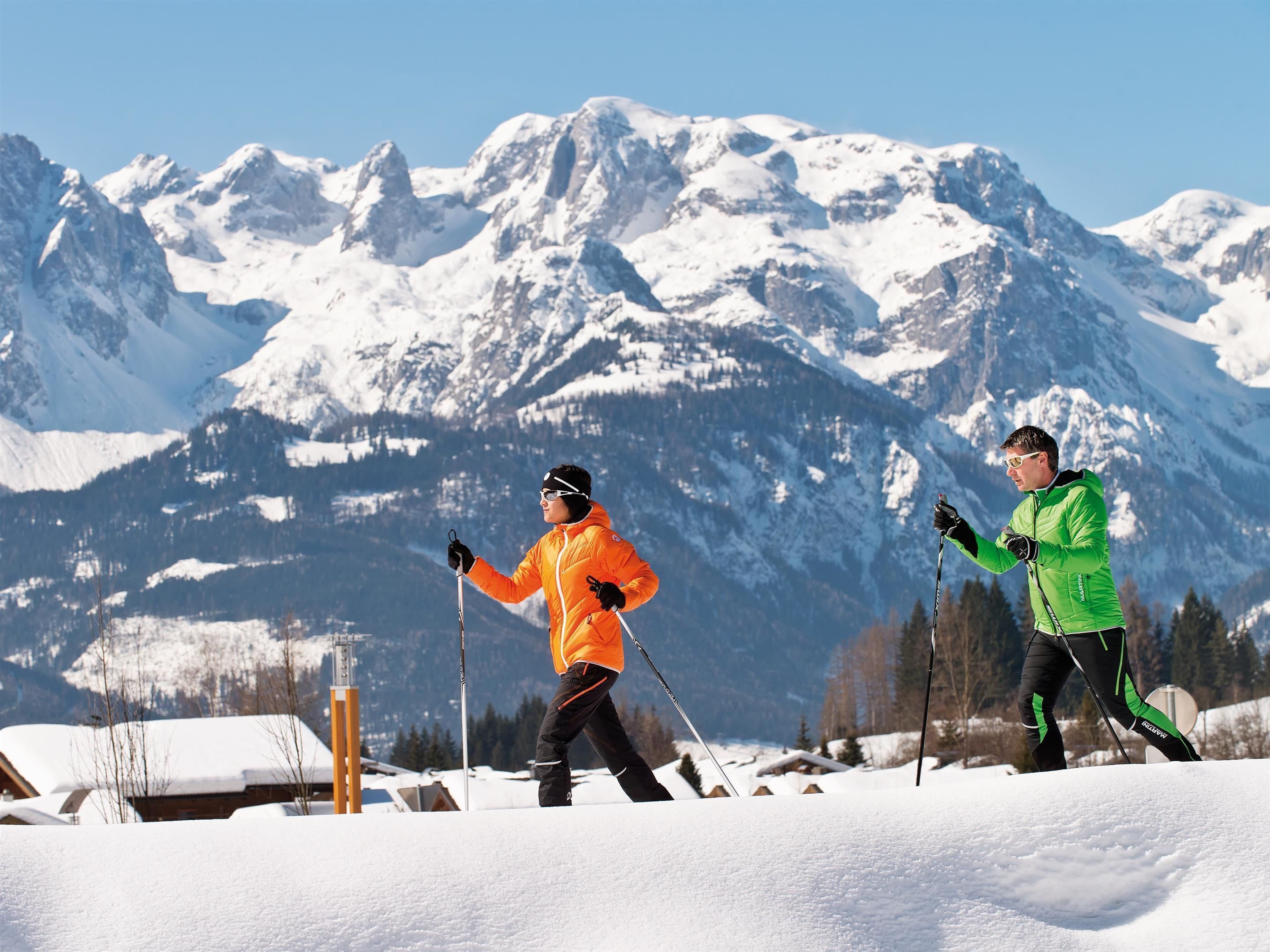 Two skiers in bright orange and green jackets running on a snowy trail with snow-covered mountains in the background.