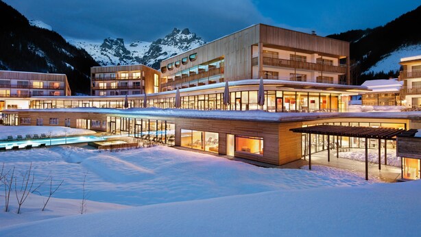 Ein modernes Hotel mit mehreren Gebäuden, einem Poolbereich und einer verschneiten Landschaft mit Bergen im Hintergrund.